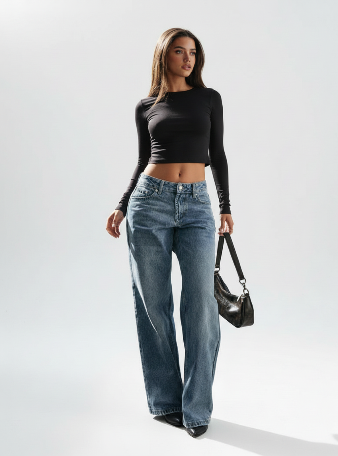 Woman wearing a black long-sleeve top and blue jeans, holding a black handbag on a white background