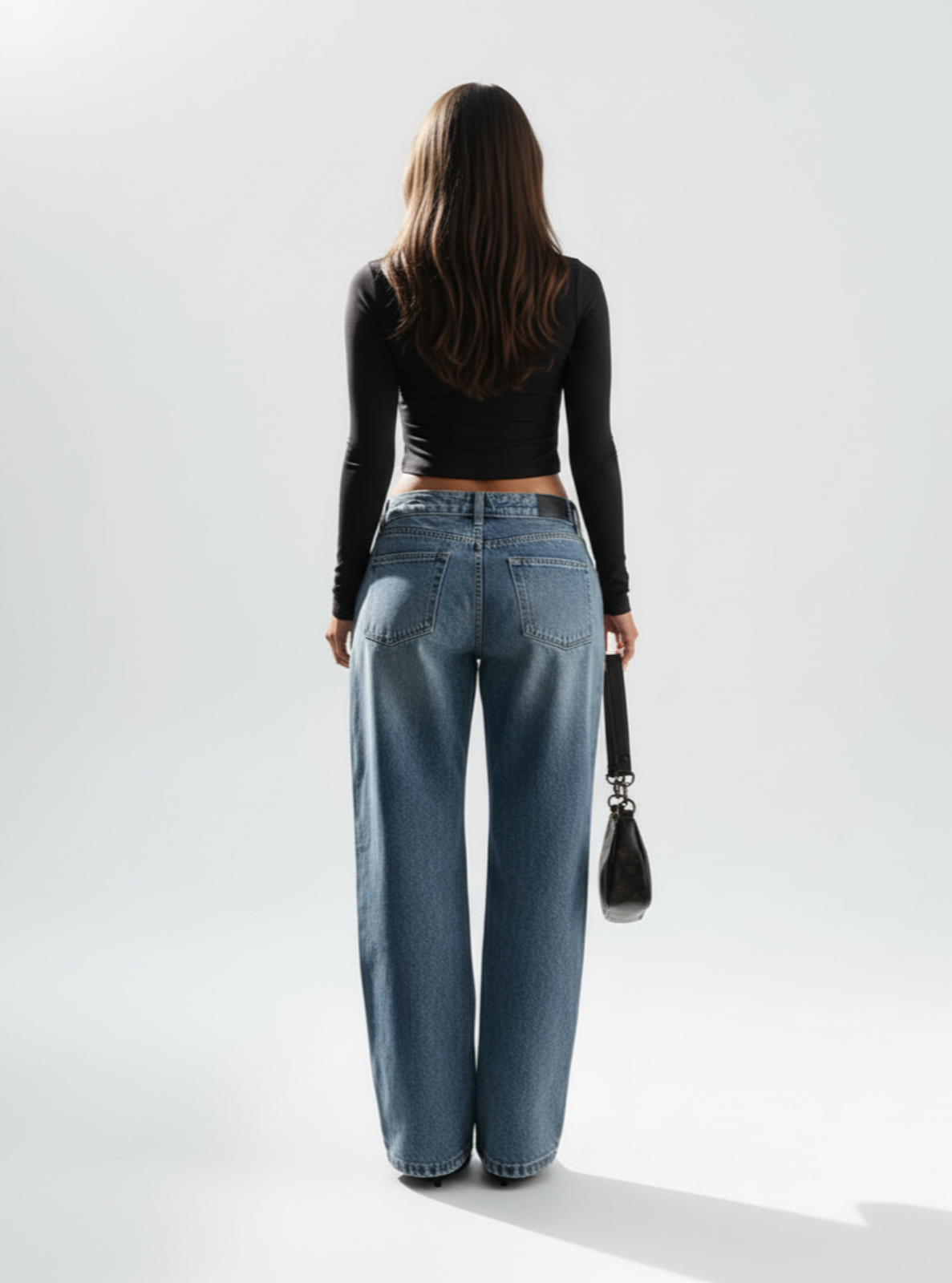 Person wearing blue jeans and a black long-sleeve top, holding a black handbag on a white background