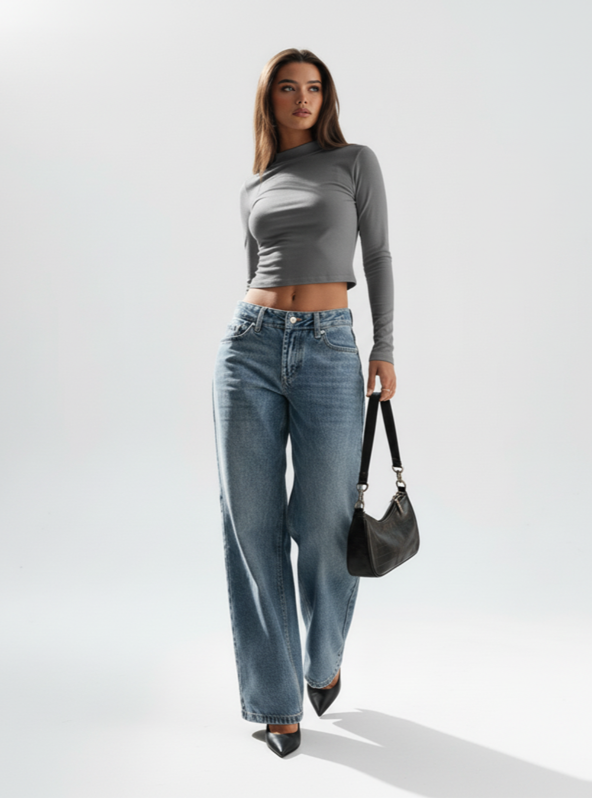 Woman wearing a gray long-sleeve top and blue jeans, holding a black handbag on a white background