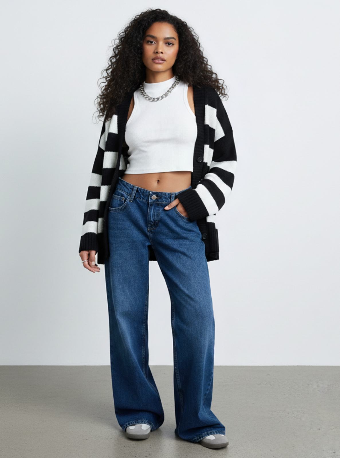 Woman wearing a black and white striped cardigan, white top, blue jeans, and silver shoes on a plain background