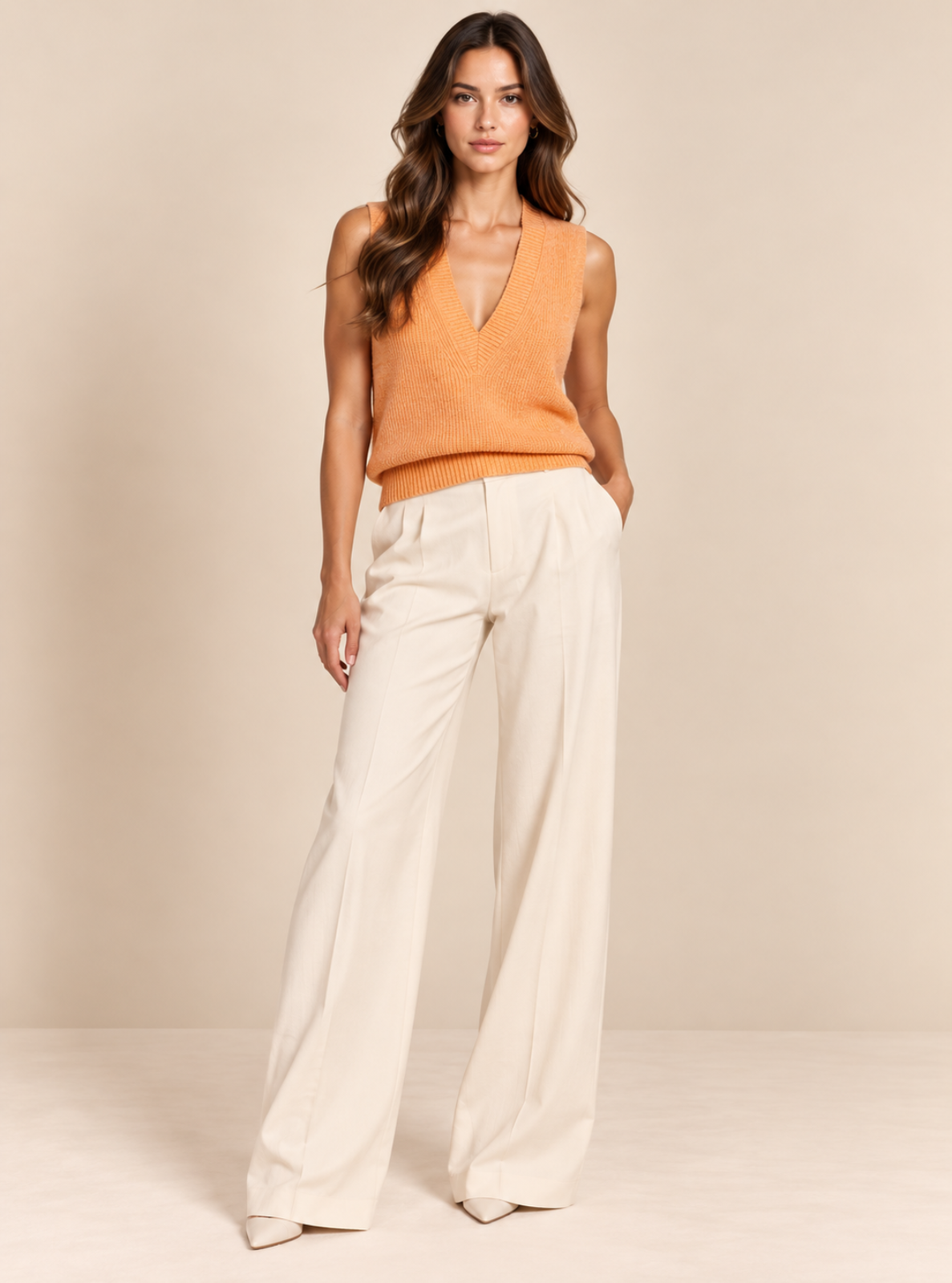 A woman modeling a sleeveless orange knit vest with a deep V-neck paired with high-waisted, wide-leg cream trousers, set against a solid beige studio backdrop.