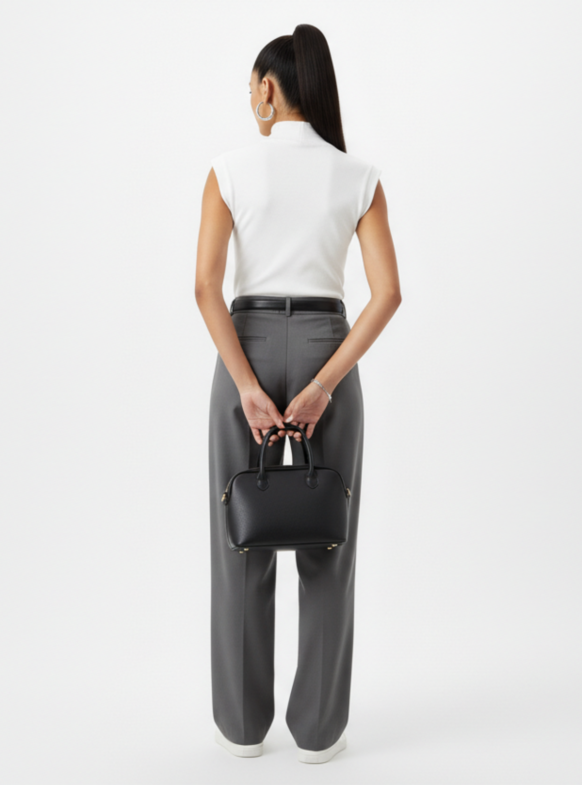 Woman holding a black handbag wearing gray pants and a white top on a white background