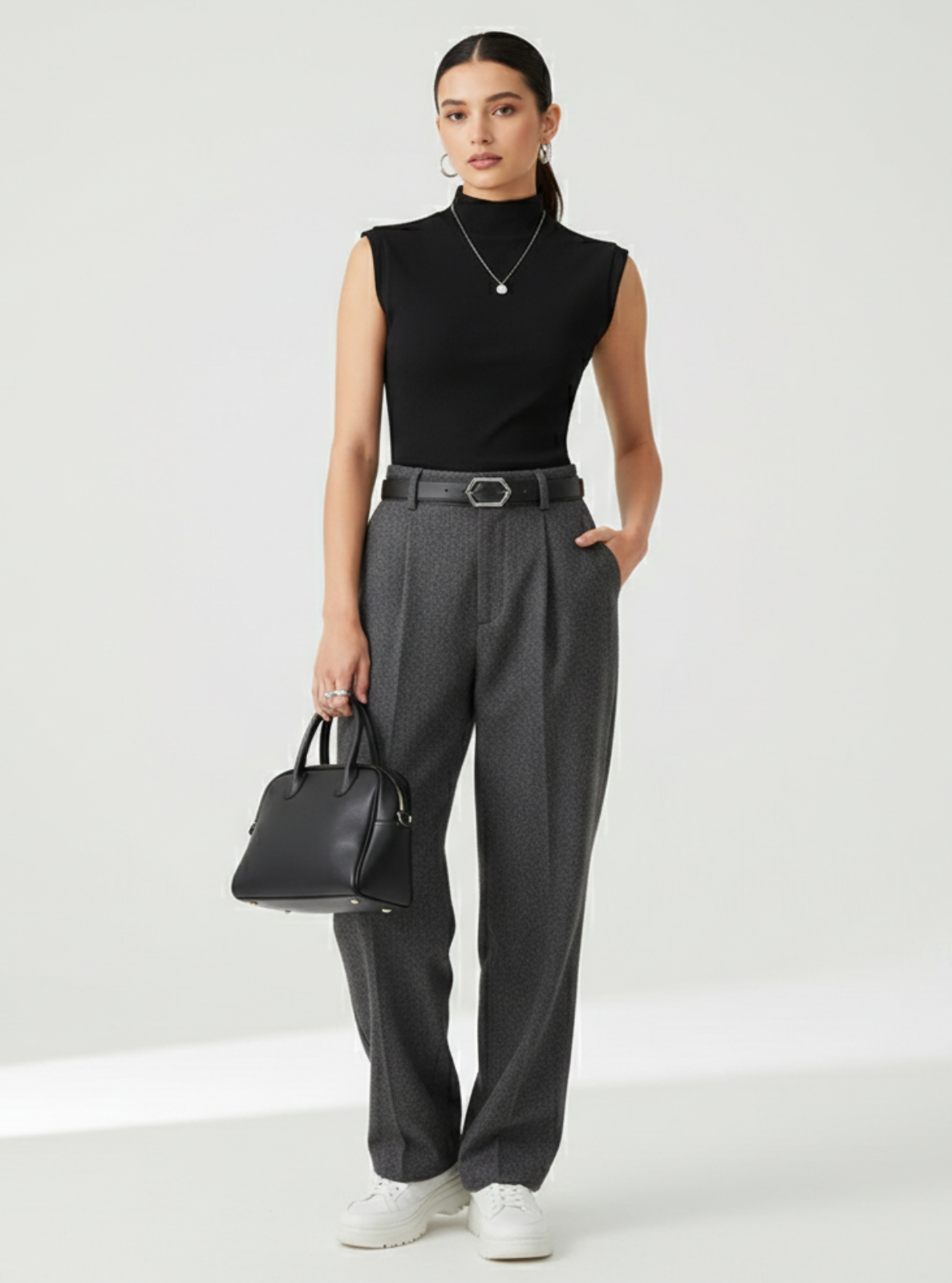 Woman wearing a black sleeveless top and gray pants, holding a black handbag on a white background