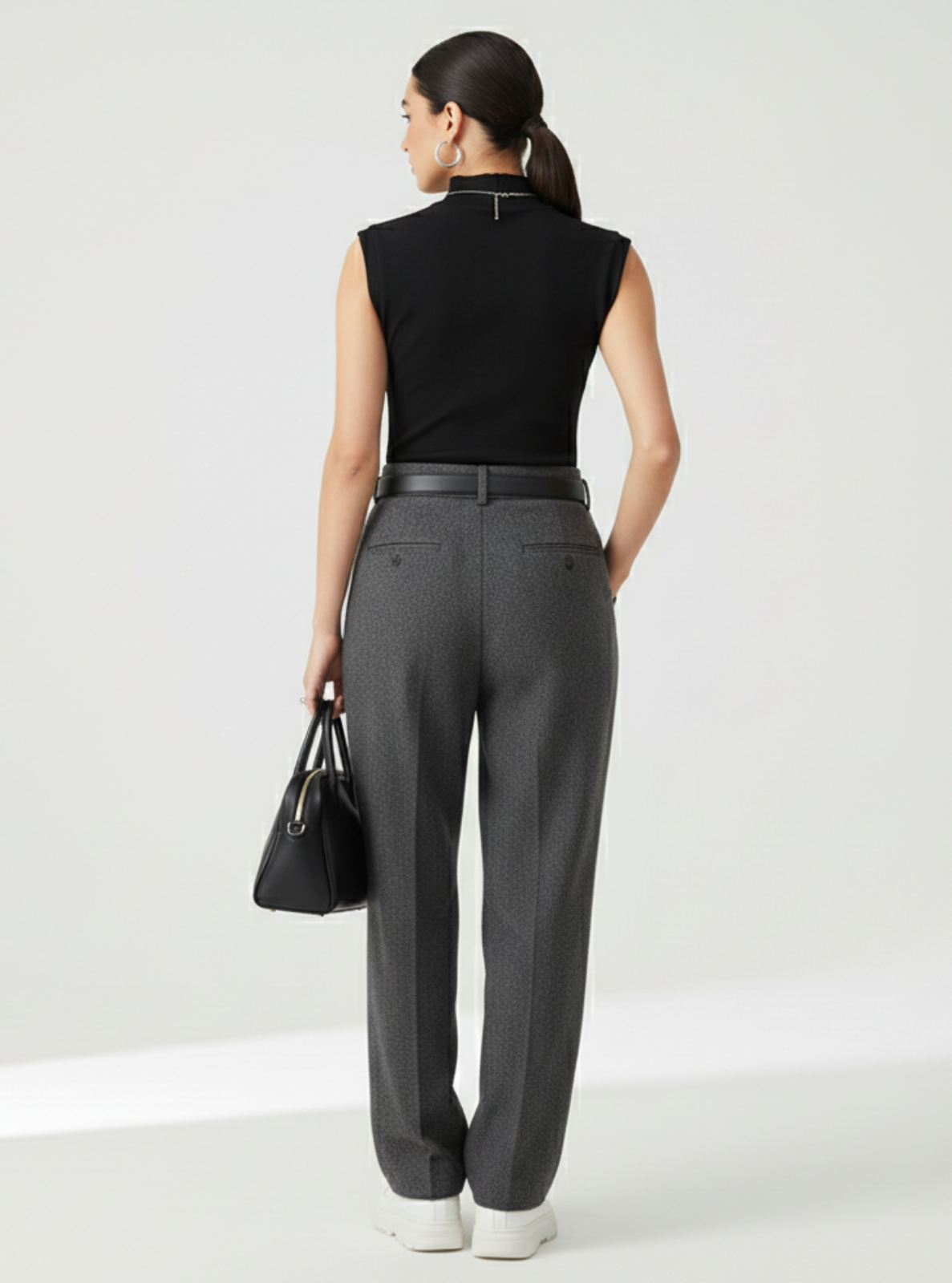 Woman wearing a black sleeveless top and gray pants, holding a black handbag on a white background