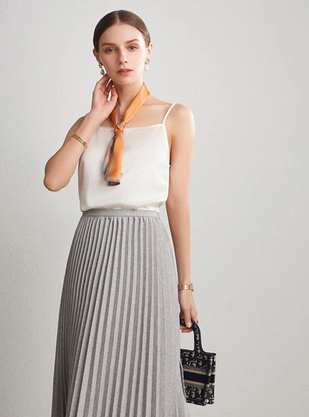 Woman wearing a white top, gray pleated skirt, and orange scarf against a plain background