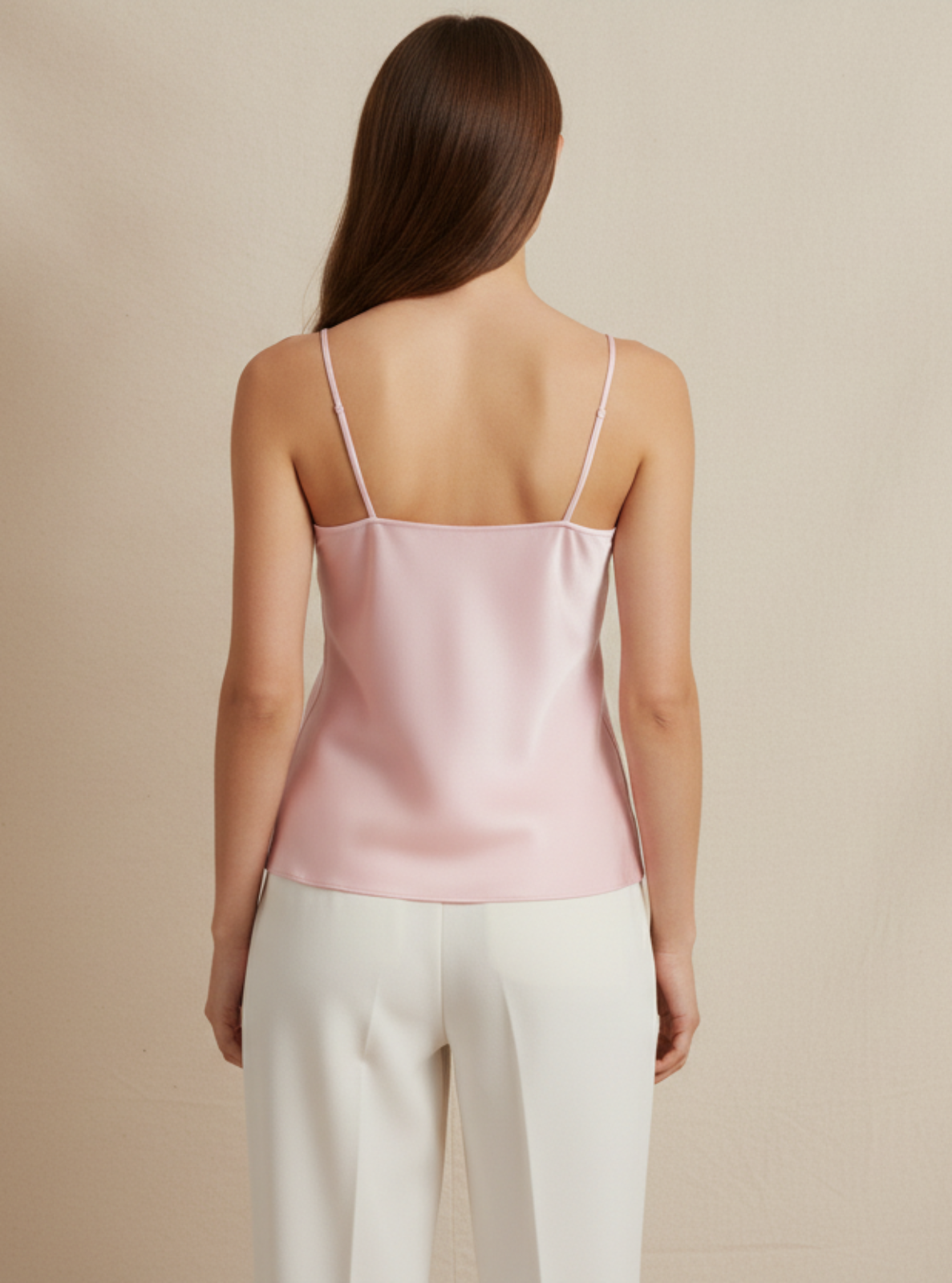 Woman wearing a pink satin camisole and white pants against a beige background