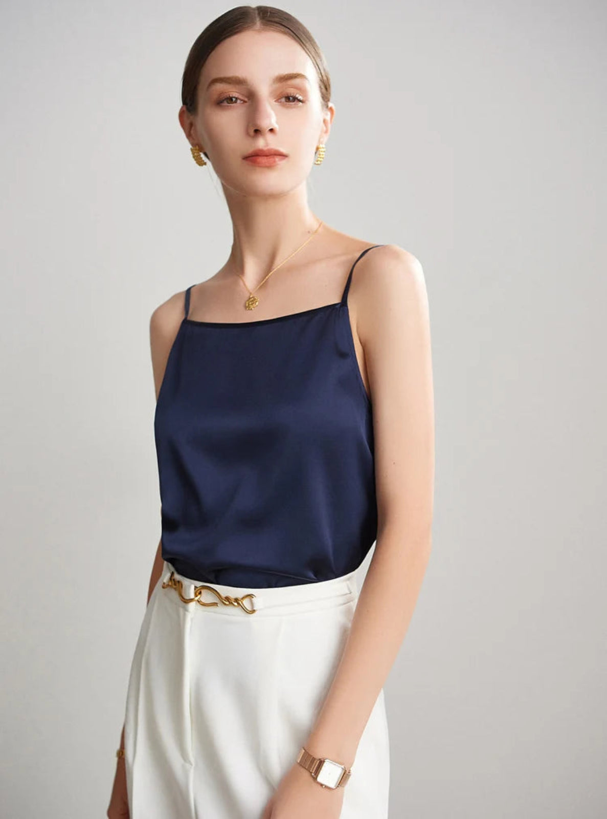 Woman wearing a navy blue satin top and white pants against a gray background