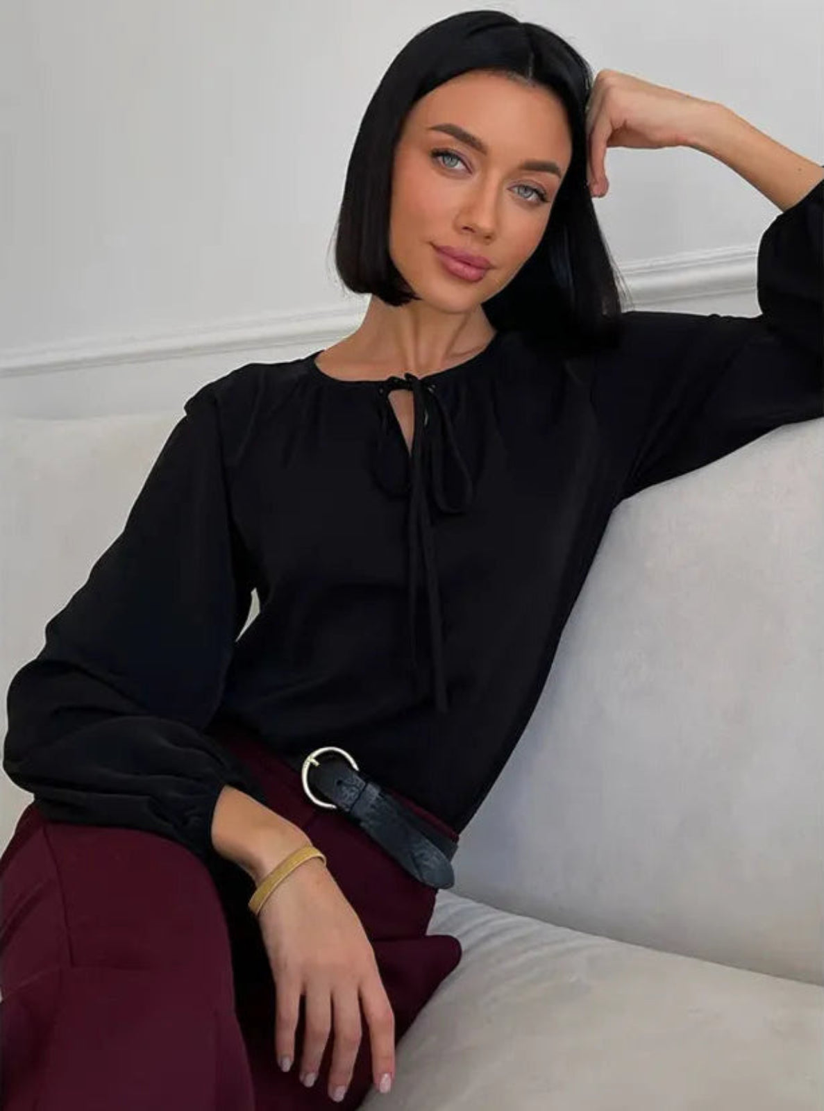 Woman in a black blouse and maroon pants sitting on a light-colored couch.