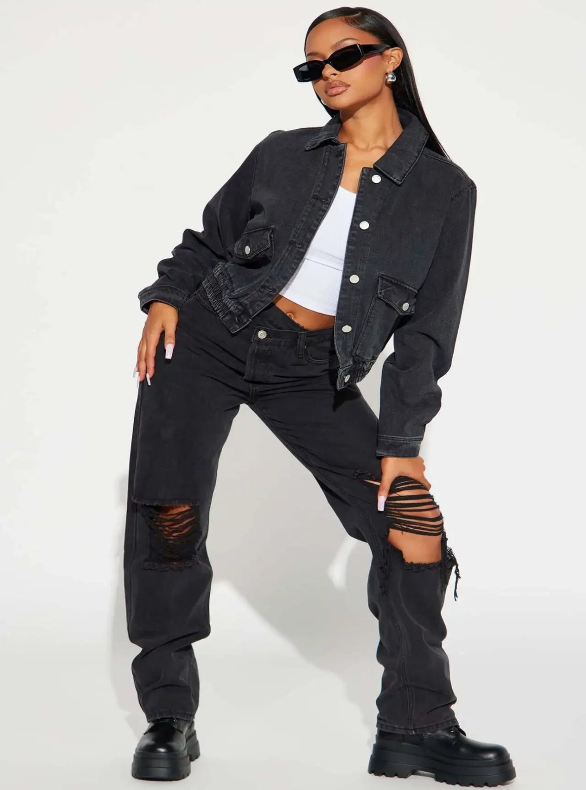 Person wearing a black denim jacket, white top, black ripped jeans, and black knee-high boots on a white background