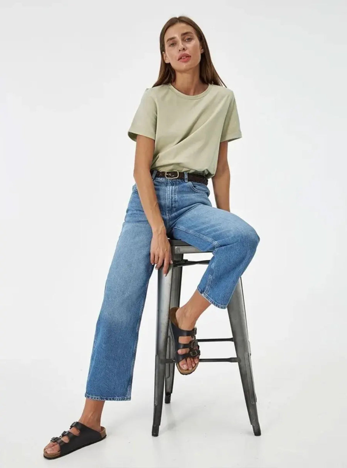 Woman sitting on a stool wearing a light green t-shirt and blue jeans against a white background