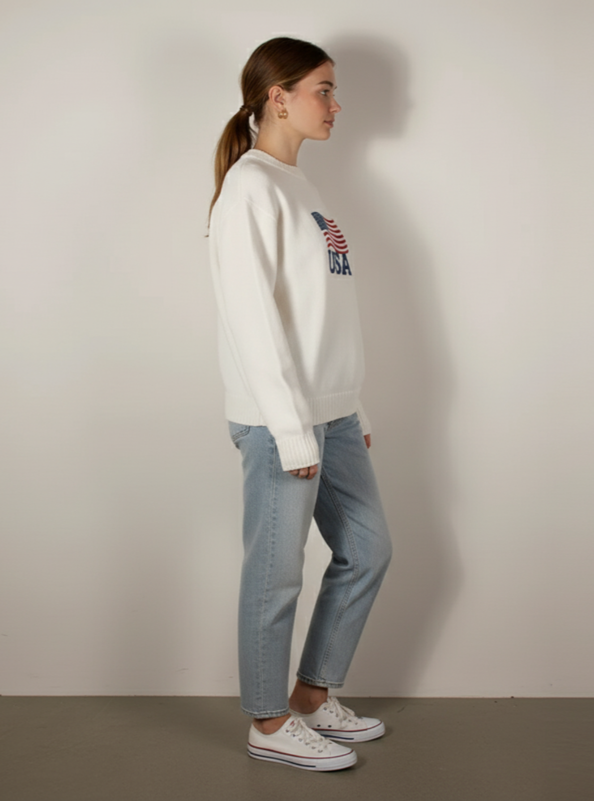 Person wearing a white sweater with a graphic design and blue jeans against a plain background