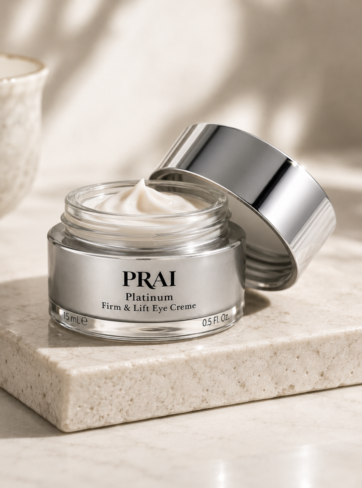 PRAI Platinum Firm & Lift Eye Crème in a clear glass jar with a reflective silver lid, shown open with cream visible on a light stone surface.