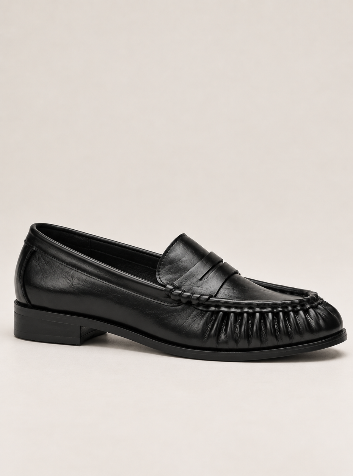 A single black leather penny loafer with a low block heel, photographed from a side profile against a plain, light beige background.