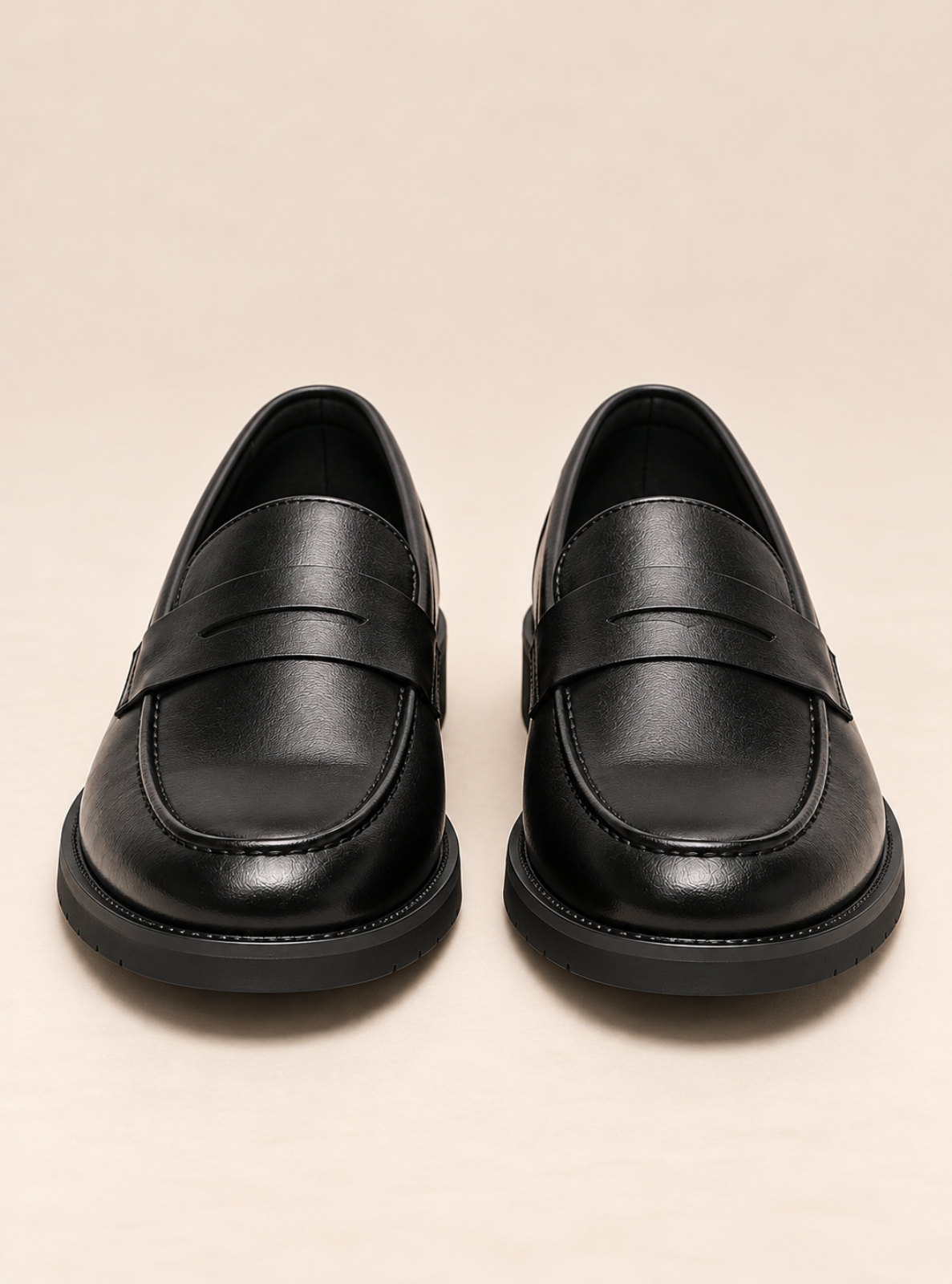 A pair of black leather penny loafers with thick black soles, photographed from a front-facing perspective on a light beige background.