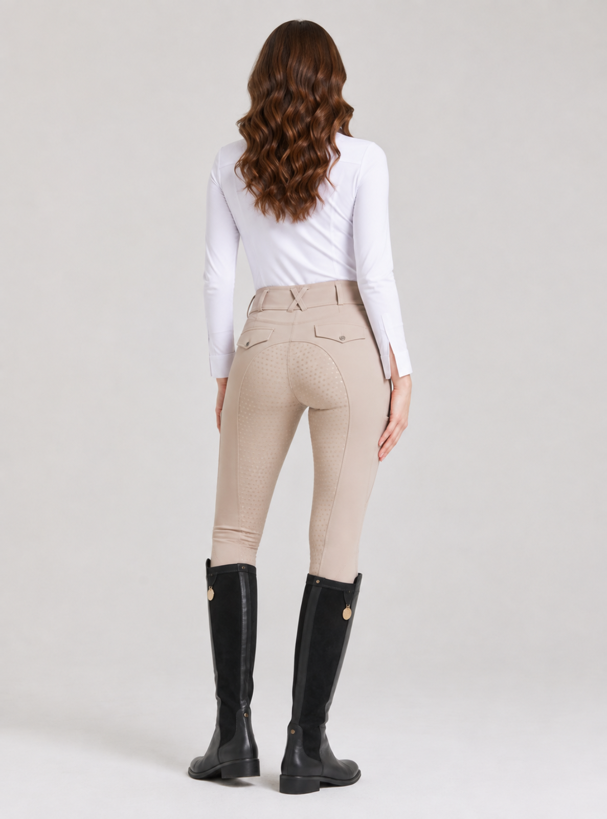 A woman shown from the back wearing beige riding breeches, a white long-sleeve top, and black riding boots against a light gray studio background.