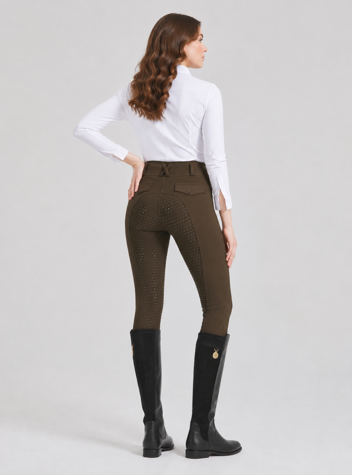 A woman shown from the back wearing high-waisted equestrian riding breeches in olive brown, a white long-sleeve shirt, and black riding boots against a light gray studio background.