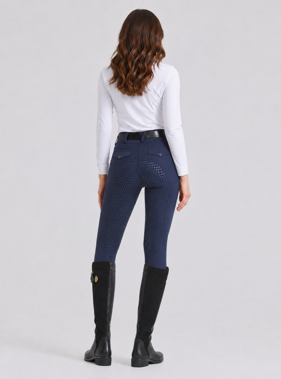 A woman viewed from the back wearing navy blue textured biker shorts, a white long-sleeve top, a black belt, and black riding boots against a light gray studio background.