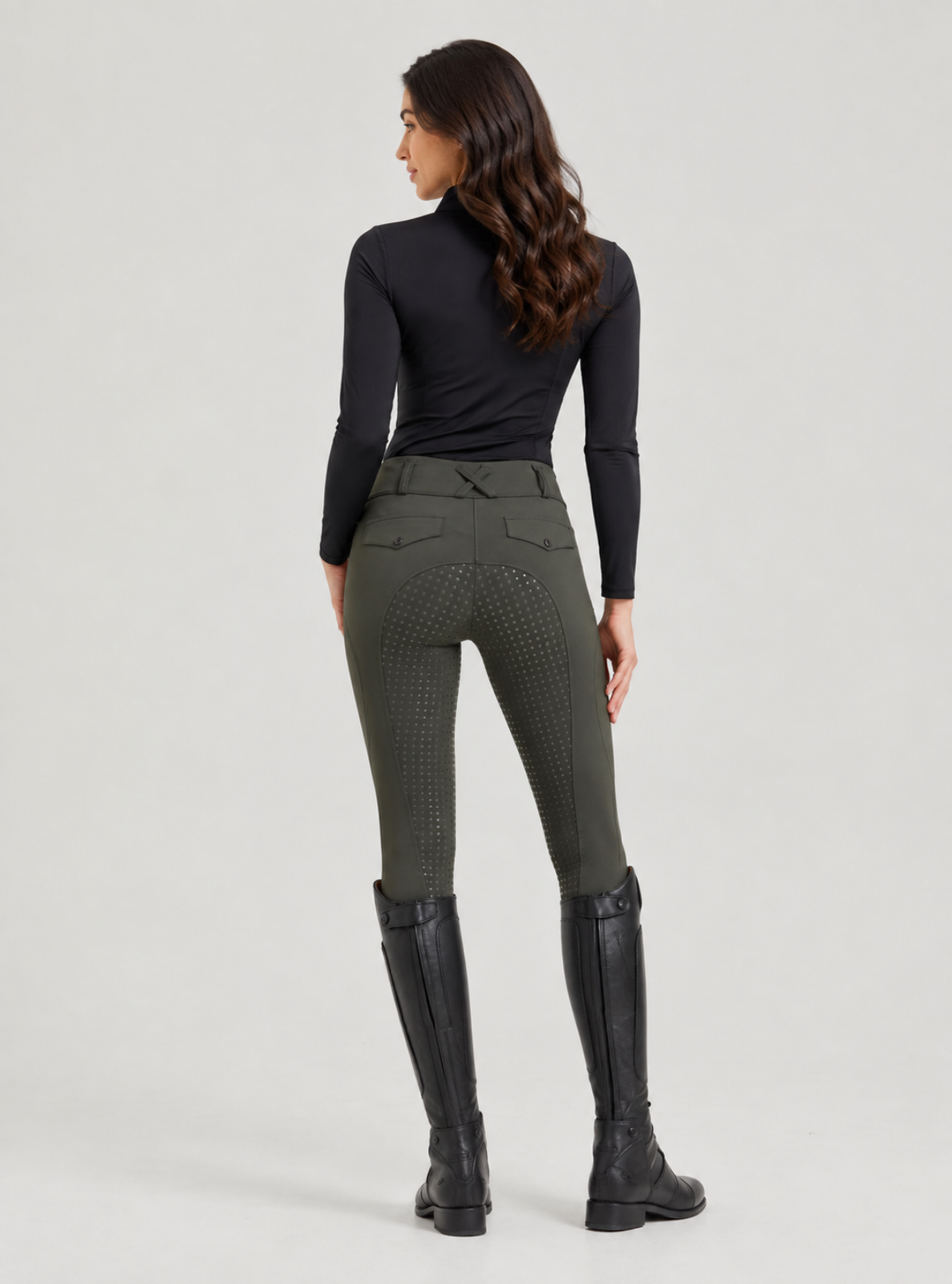 A woman shown from the back wearing fitted dark olive riding breeches with a textured pattern, a black long-sleeve top, and black riding boots against a light gray studio background.
