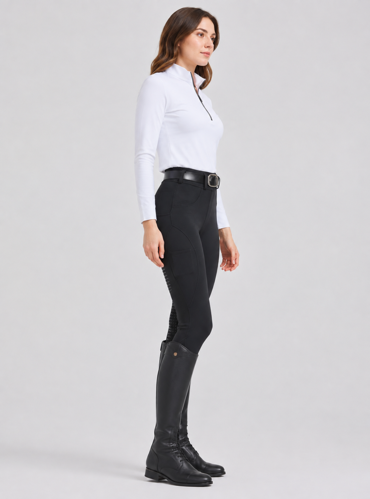 A woman in side profile wearing a white long-sleeve riding shirt, black equestrian breeches with knee patches, a black belt, and black riding boots, photographed against a light gray studio background.