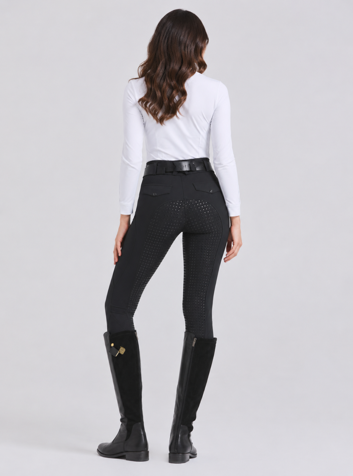 A woman shown from the back wearing black equestrian breeches with a textured pattern, a white long-sleeve shirt, a black belt, and black riding boots against a light gray studio background.