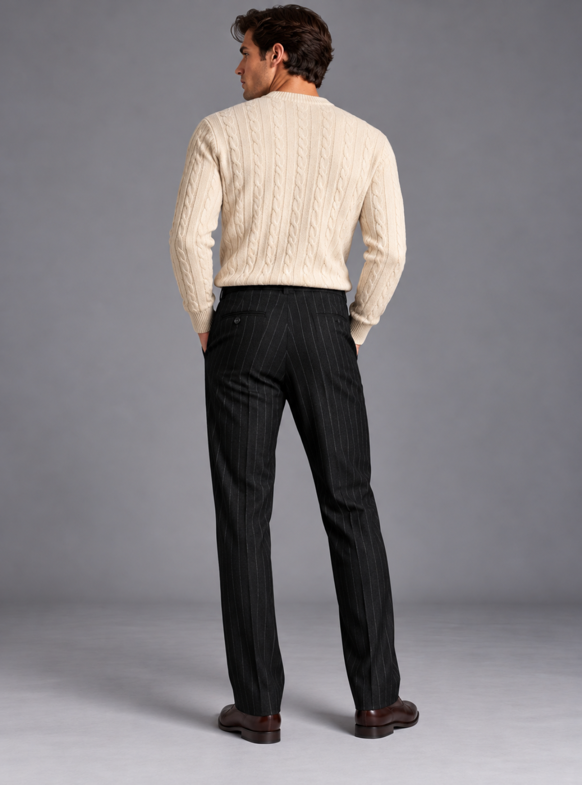 A man viewed from behind wearing a cream cable-knit sweater and black pinstripe trousers, standing against a neutral gray studio background.