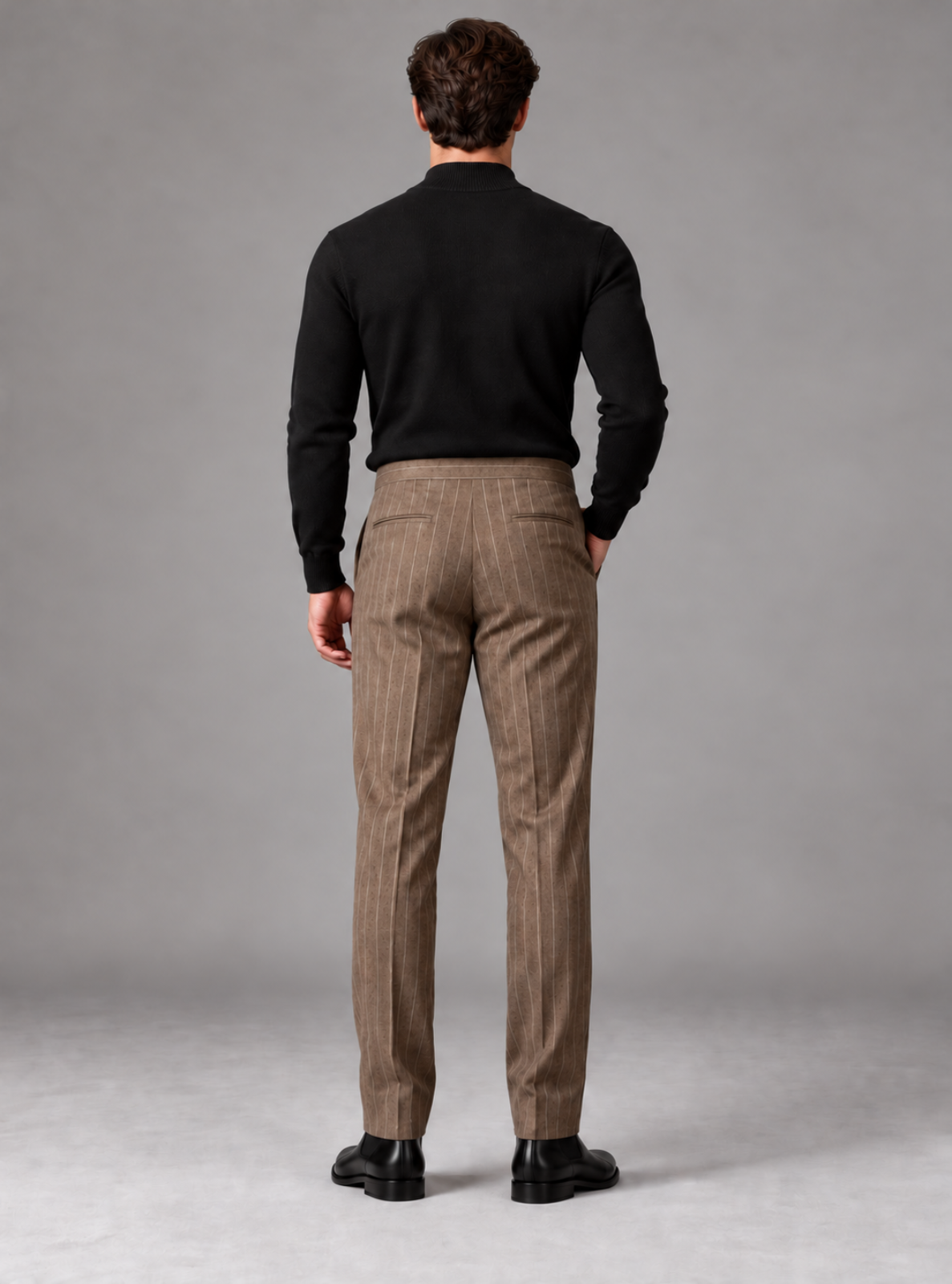 A man viewed from behind wearing brown pinstripe trousers, a black long-sleeve shirt, and black dress shoes, standing against a neutral gray studio background.