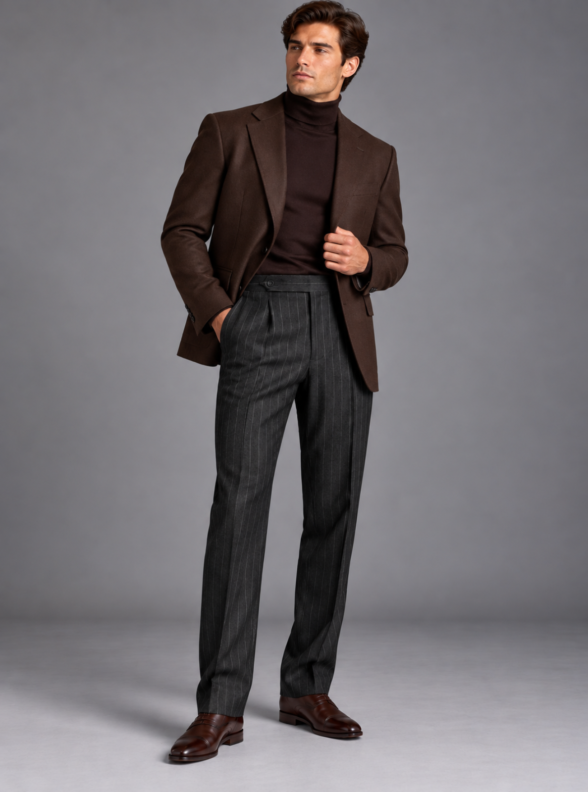 A man modeling a dark brown blazer over a dark brown turtleneck sweater, paired with dark gray pinstriped trousers and brown leather dress shoes, against a solid gray studio background.