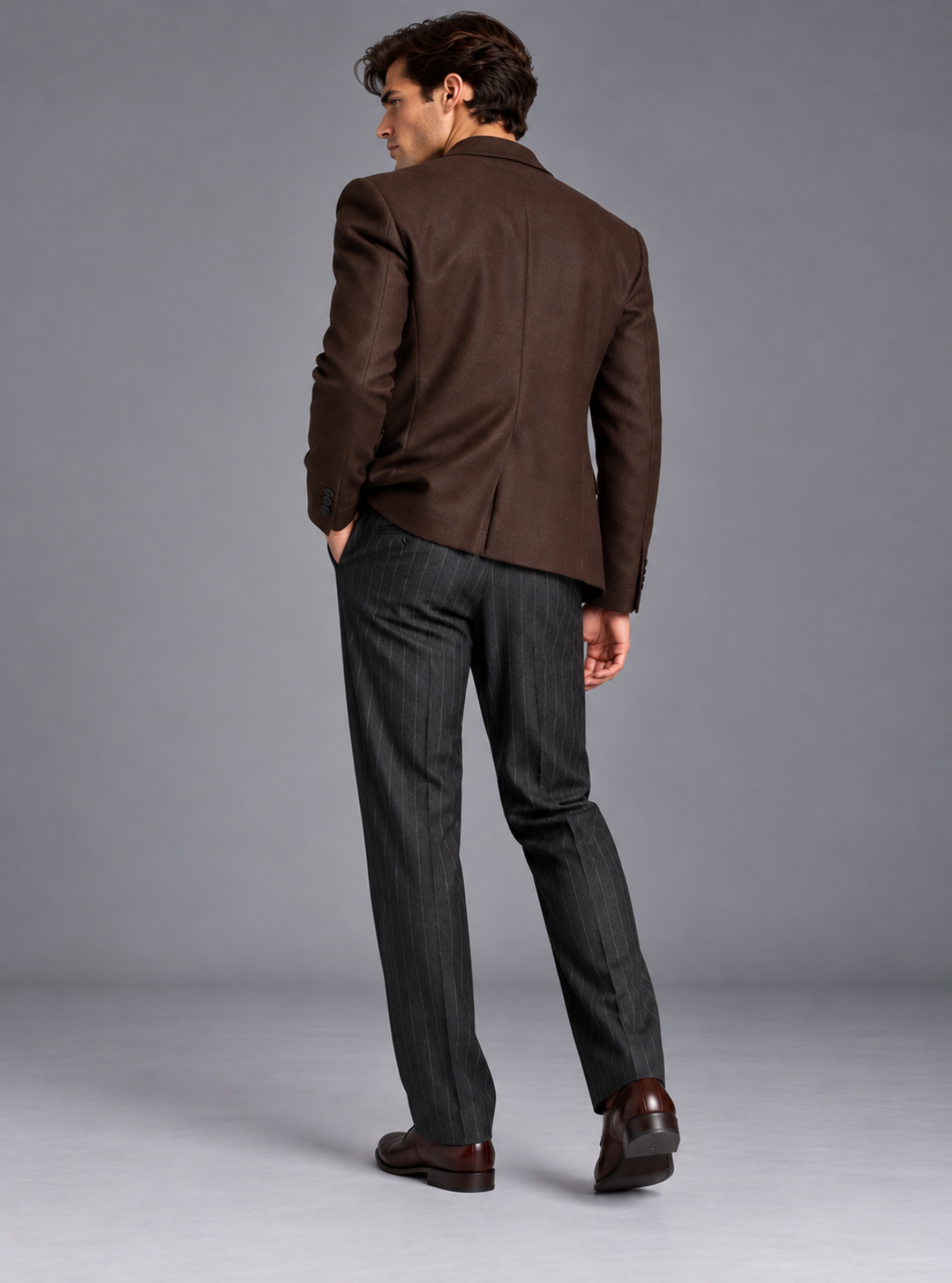 A man wearing a brown blazer and dark gray pinstriped trousers, shown from behind in a studio setting with a gray background.
