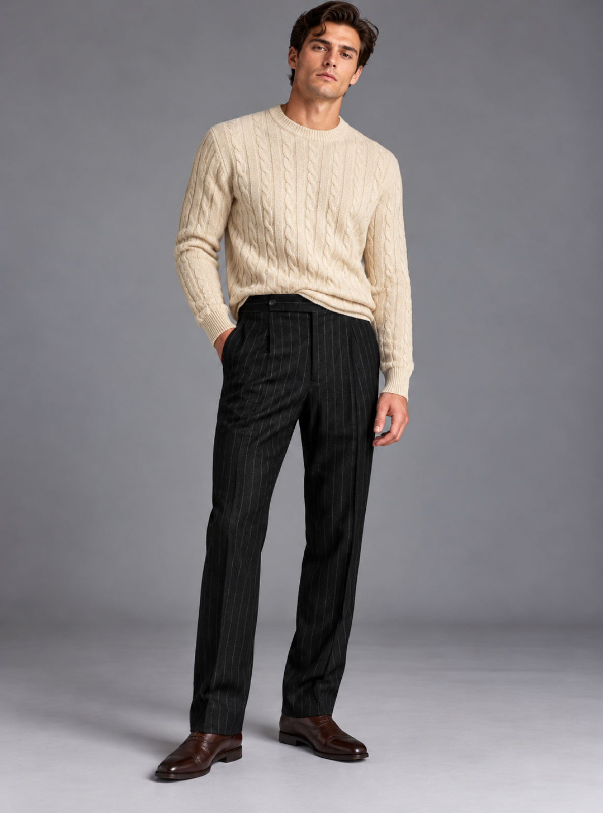 A man modeling a cream cable-knit crewneck sweater and black pinstriped trousers with brown leather dress shoes, standing against a solid gray studio background.