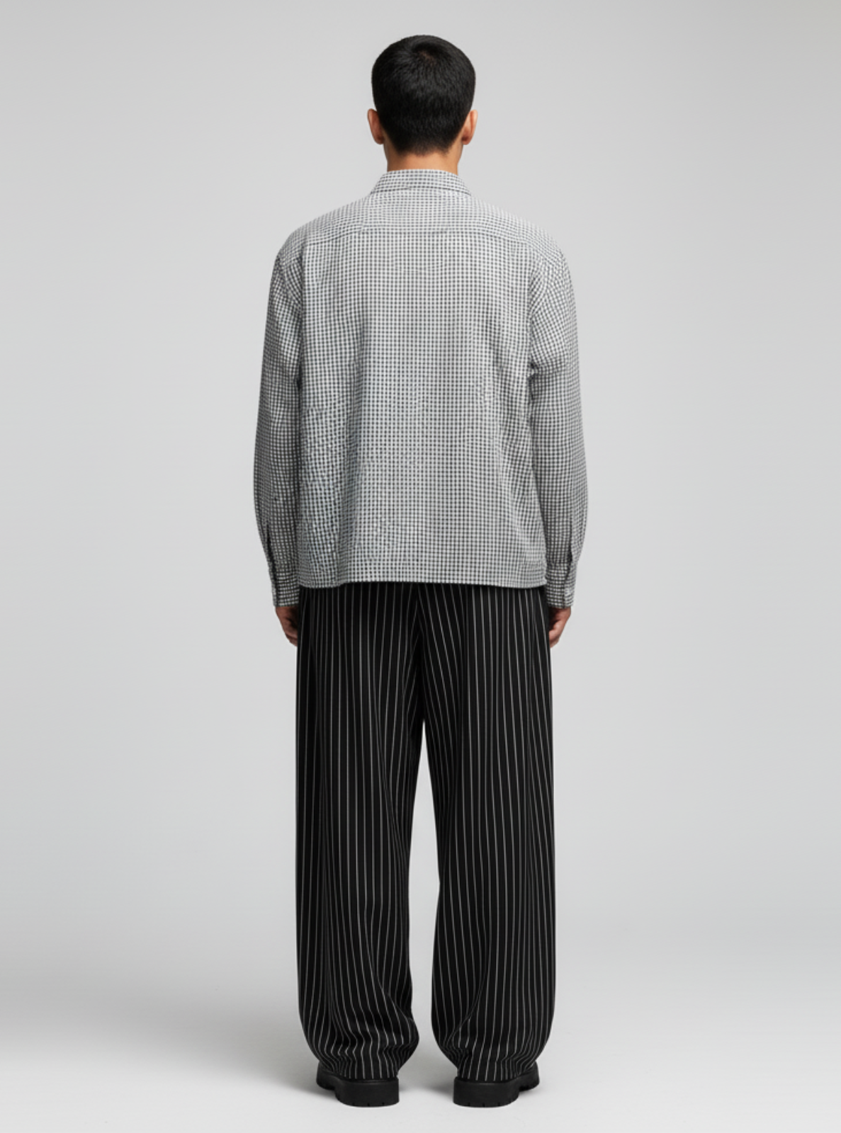 Person wearing a checkered shirt and black pleated pants on a gray background