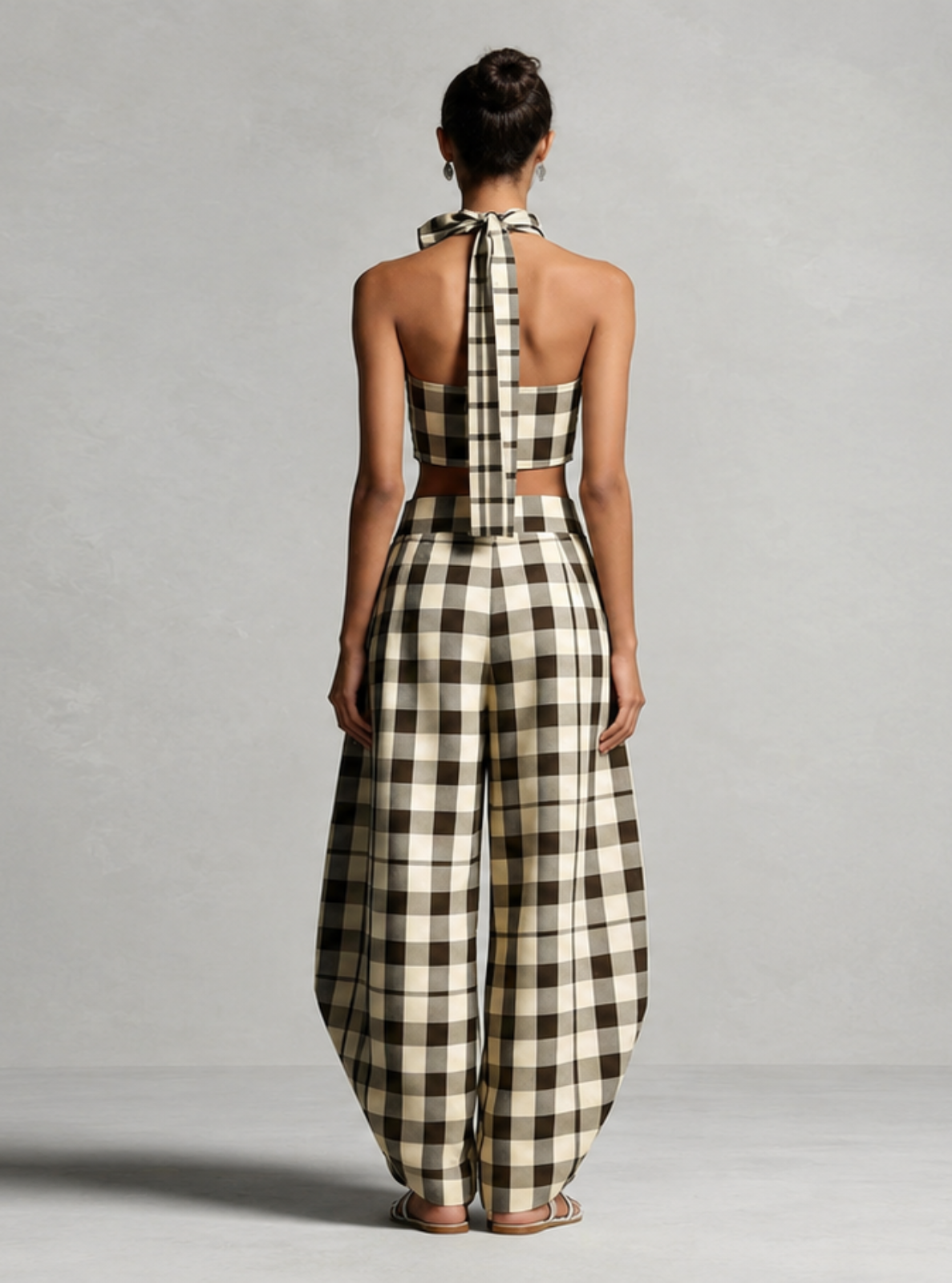 A model viewed from behind wearing a beige and black gingham halter-neck jumpsuit with a cutout at the waist, standing against a neutral gray studio background.