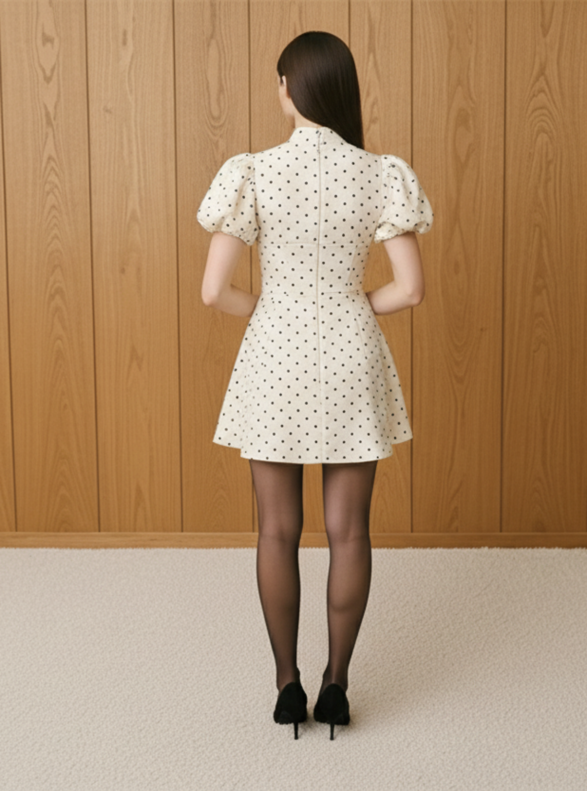 Woman wearing a white polka dot dress standing against a wooden paneled wall.
