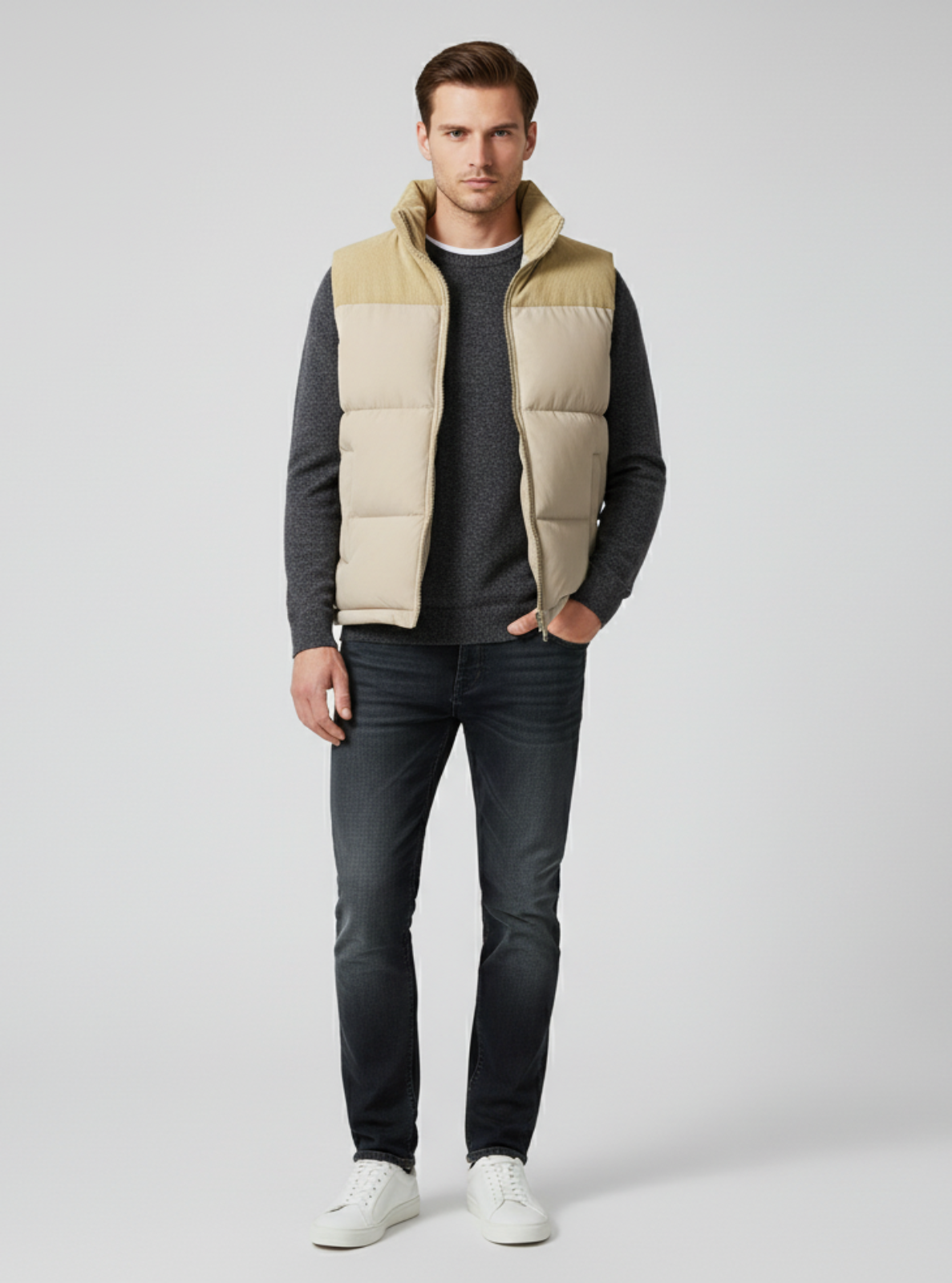 Man wearing a beige and brown puffer vest over a dark long-sleeve shirt, black jeans, and white sneakers on a light gray background.
