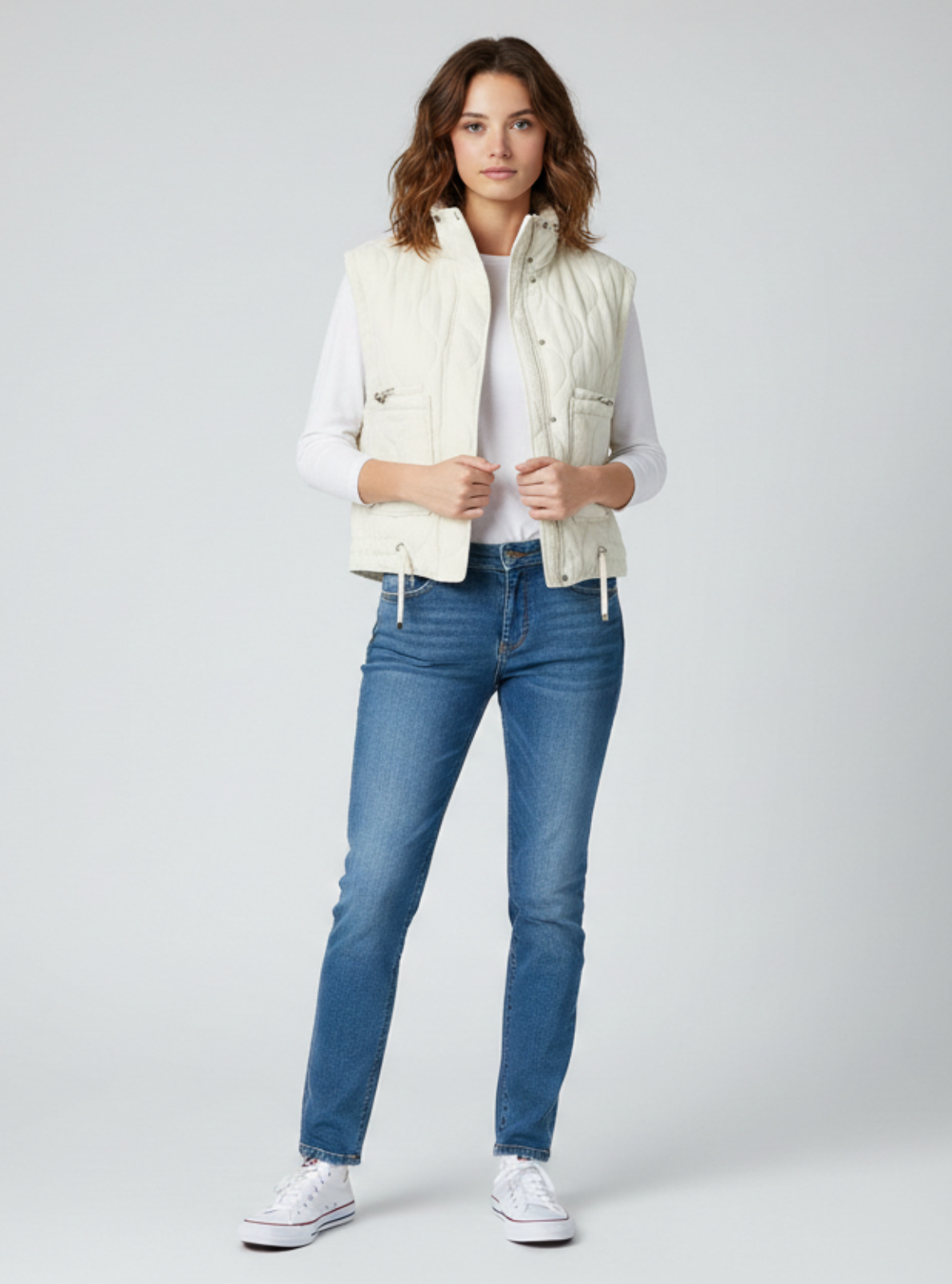 Woman wearing a white vest, white shirt, blue jeans, and white sneakers on a light gray background