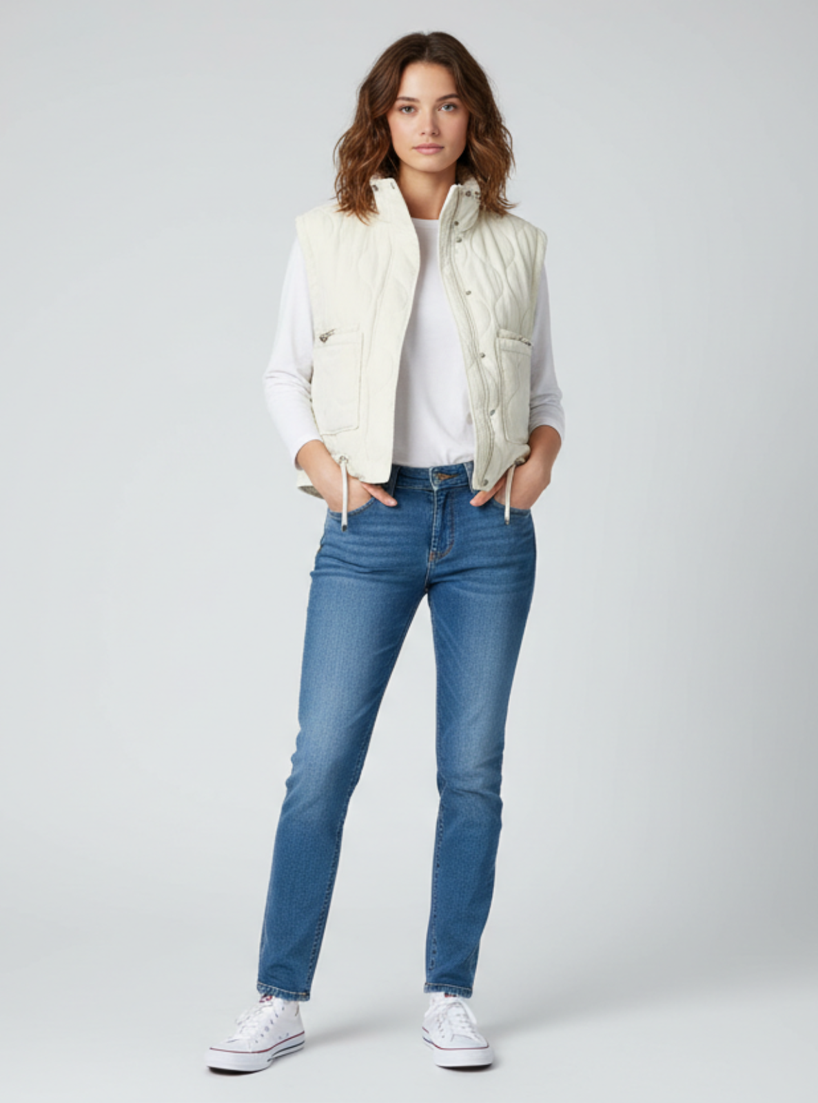 Woman wearing a white puffer vest, white shirt, blue jeans, and white sneakers on a light gray background