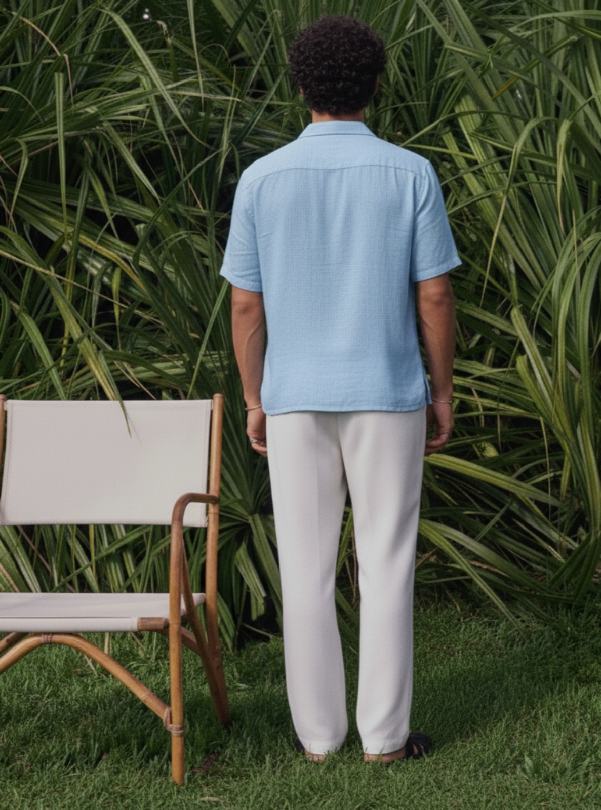 Person wearing a light blue shirt and white pants standing in a garden with green foliage.