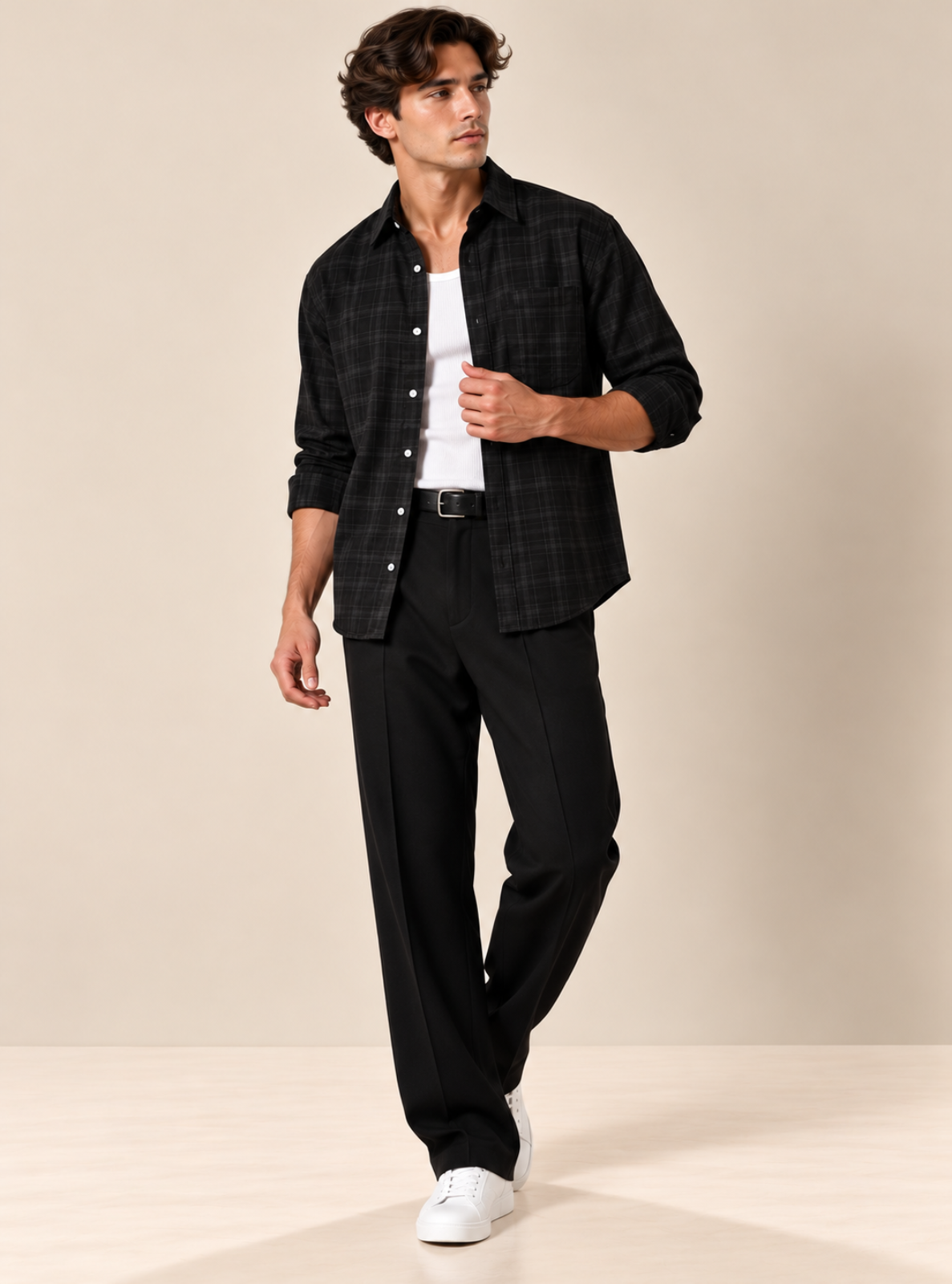 A man modeling a black plaid button-down shirt over a white t-shirt, paired with black trousers and white sneakers, against a solid beige studio background.
