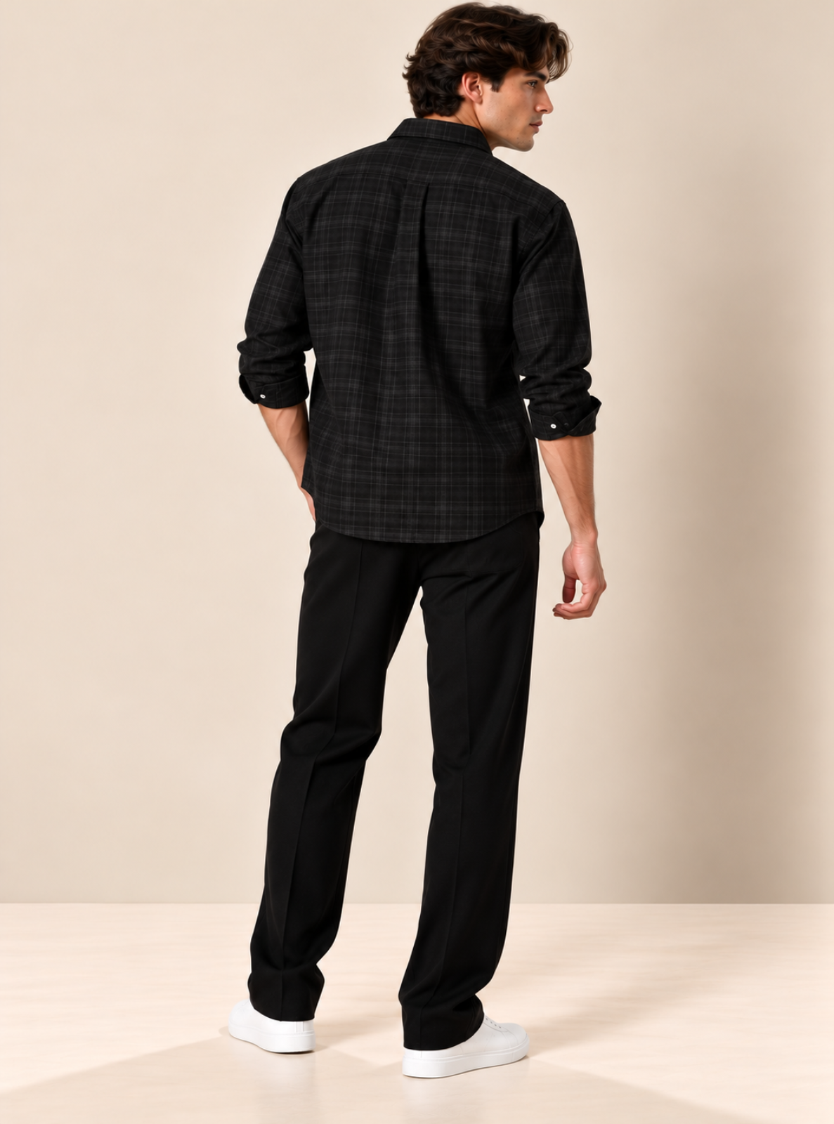 A man wearing a black and dark gray plaid button-down shirt, black trousers, and white sneakers, shown from a rear three-quarter view against a warm beige studio background.