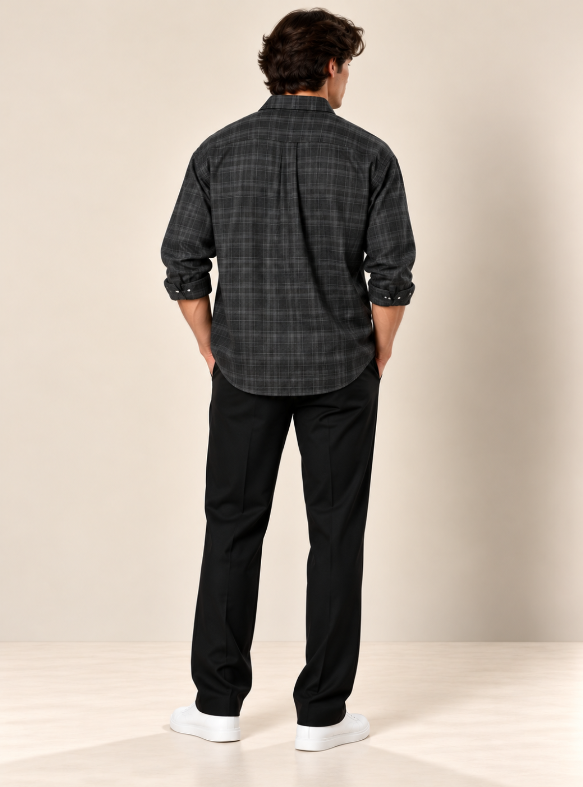 A man viewed from behind wearing a dark gray and black plaid button-down shirt, black trousers, and white sneakers, standing against a plain beige studio background.