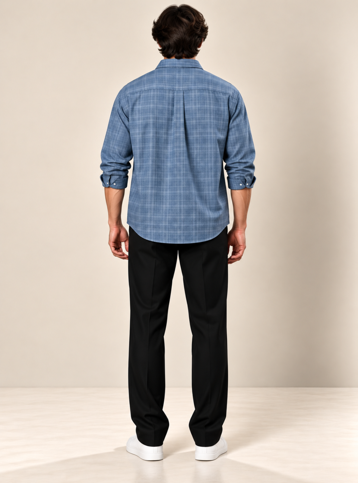 A man viewed from behind wearing a blue plaid button-down shirt, black trousers, and white sneakers, standing against a neutral beige studio background.