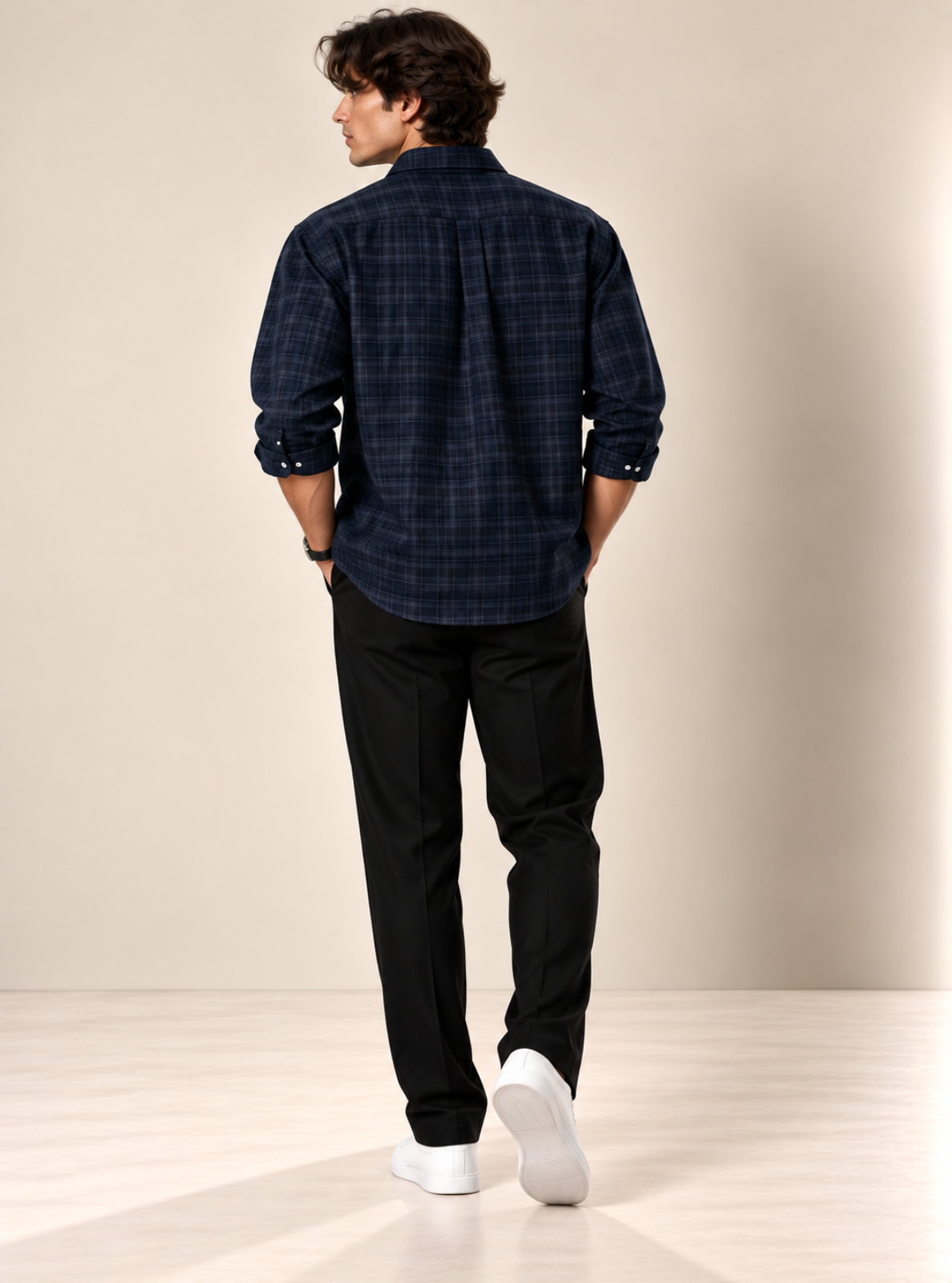 A man shown from the back wearing a dark navy blue plaid button-down shirt, black trousers, and white sneakers, standing in a studio with a beige background.