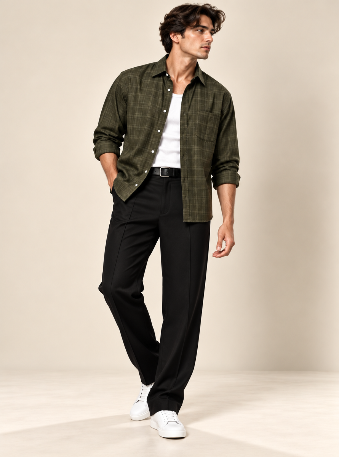 A man modeling a dark green plaid button-down shirt over a white t-shirt, paired with black trousers and white sneakers, against a neutral beige studio background.