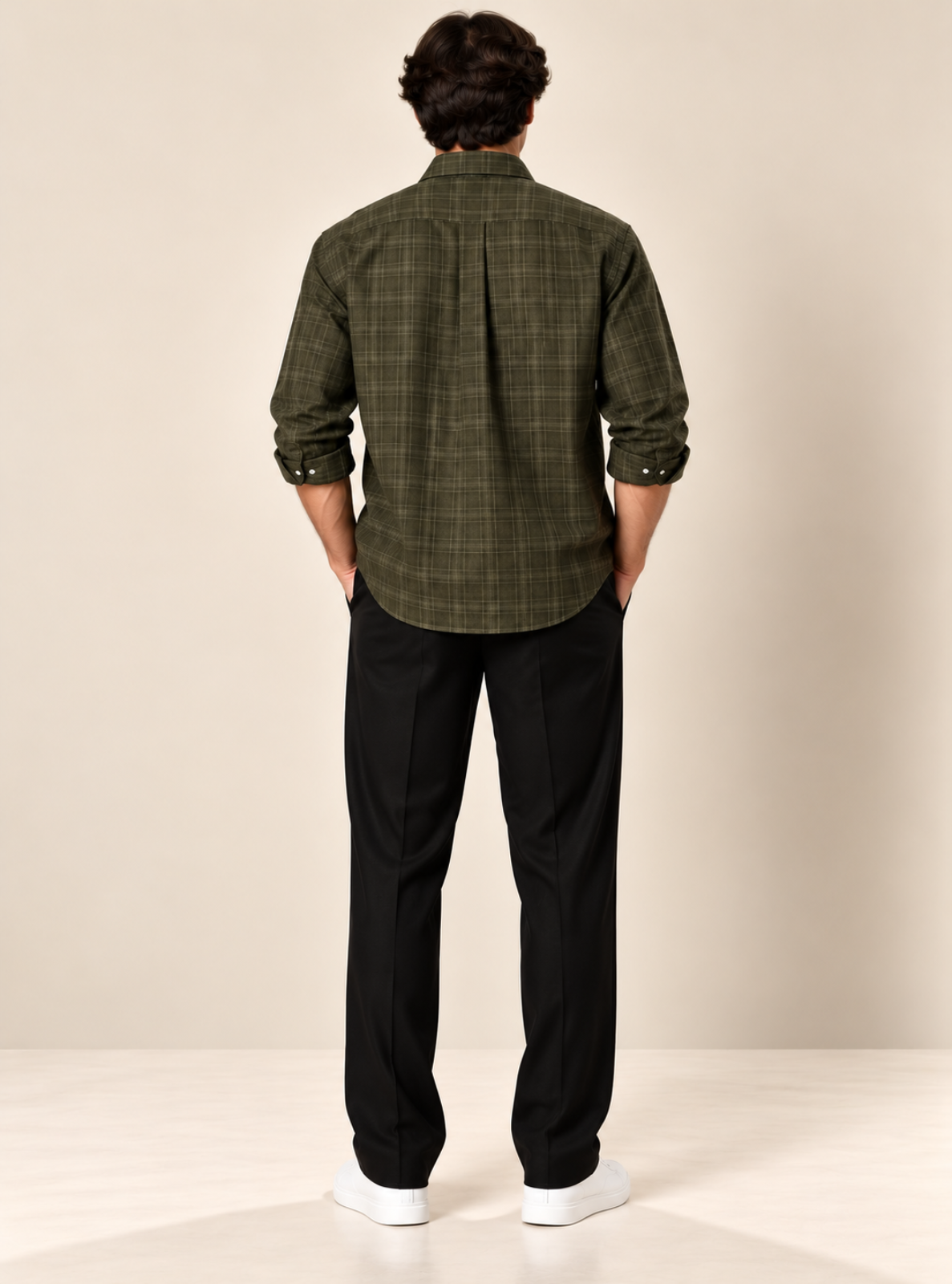 A man viewed from behind wearing an olive green plaid button-down shirt, black trousers, and white sneakers, standing against a neutral beige studio background.