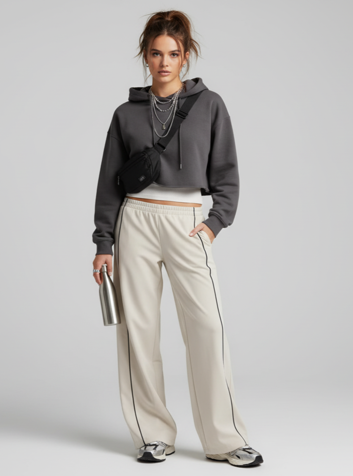 Woman wearing a gray hoodie and beige pants holding a silver water bottle on a plain background