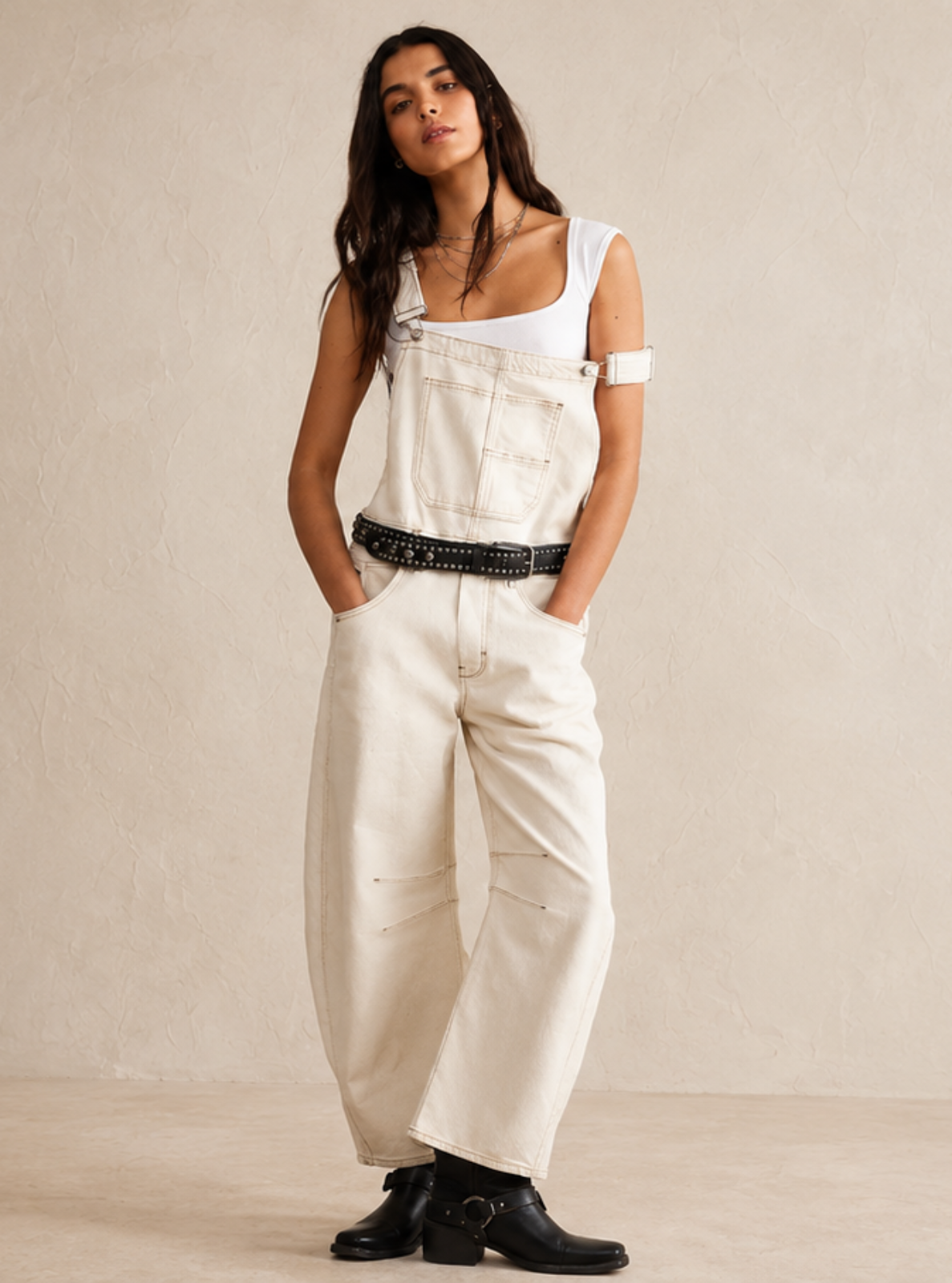 A woman modeling a pair of cream-colored denim overalls with a black leather belt, styled with black ankle boots, standing against a textured beige plaster wall.