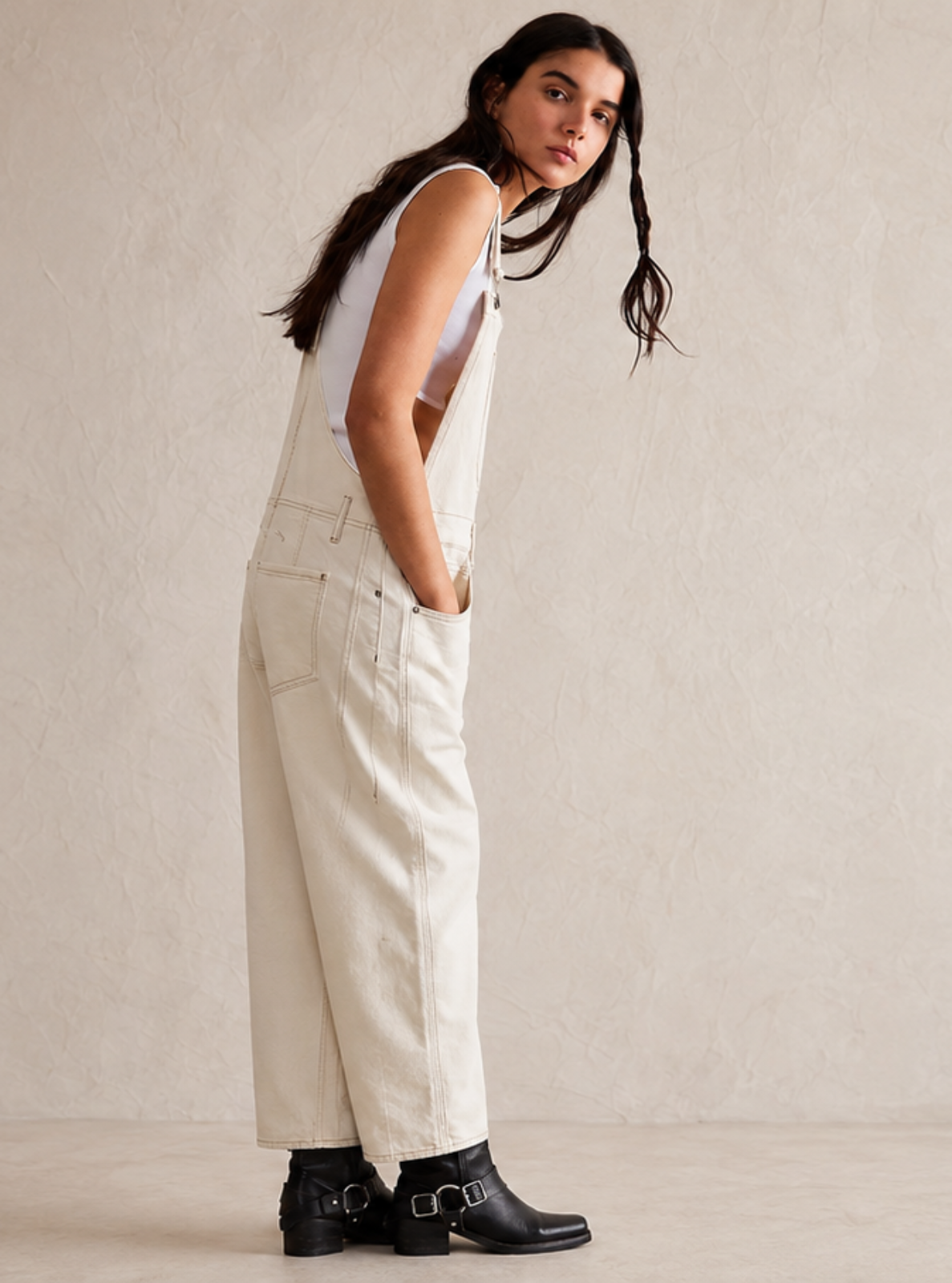 A woman modeling a sleeveless white overall-style jumpsuit with wide legs, paired with black ankle boots, standing against a neutral textured beige studio wall.