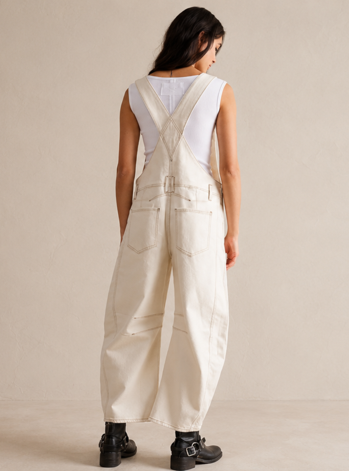 A woman viewed from behind wearing a cream-colored sleeveless overall jumpsuit with wide legs and black boots, standing against a neutral beige studio background.