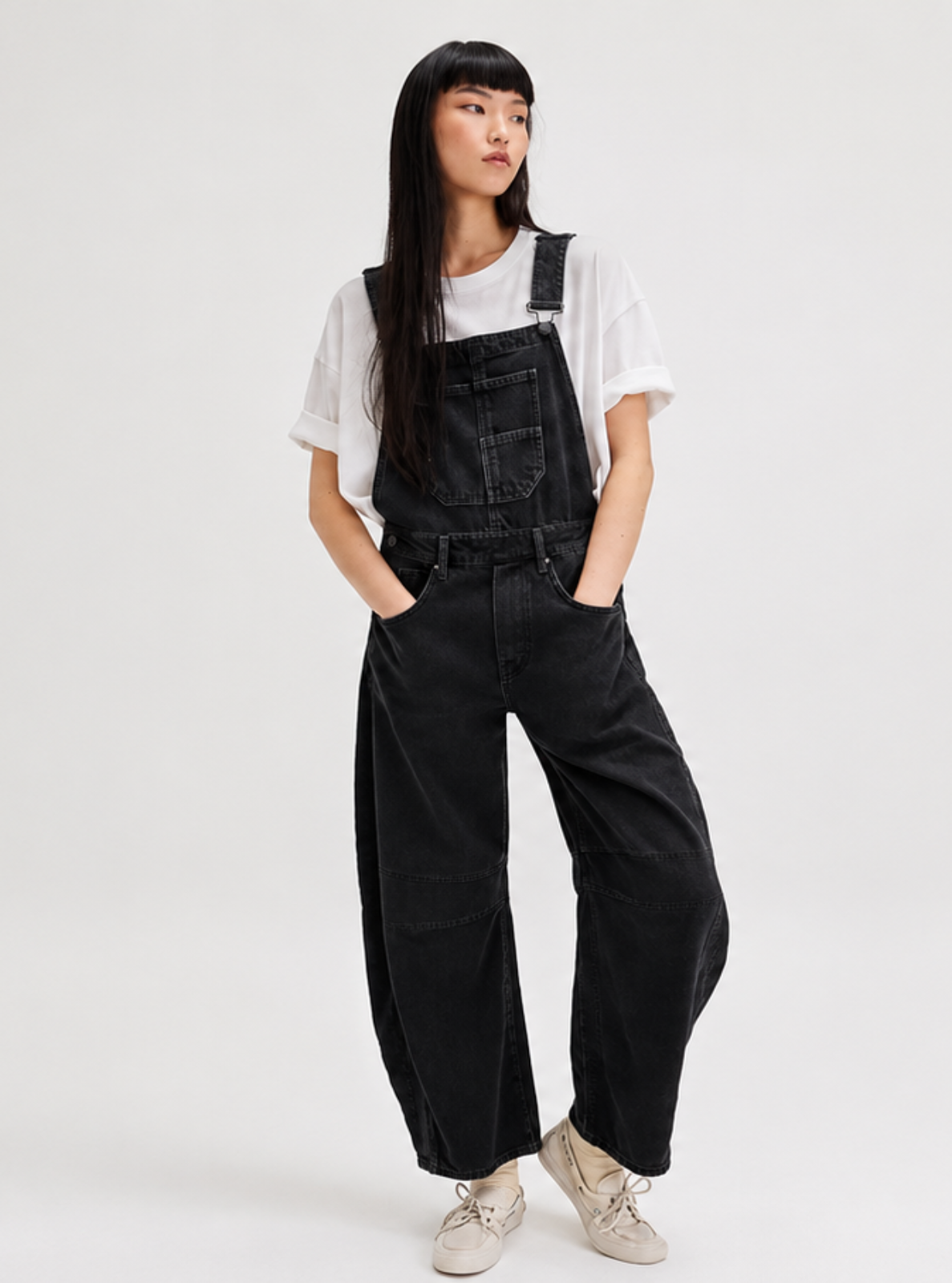 A model wearing black wide-leg denim overalls over a white short-sleeve t-shirt and white sneakers, standing against a plain light gray studio background.
