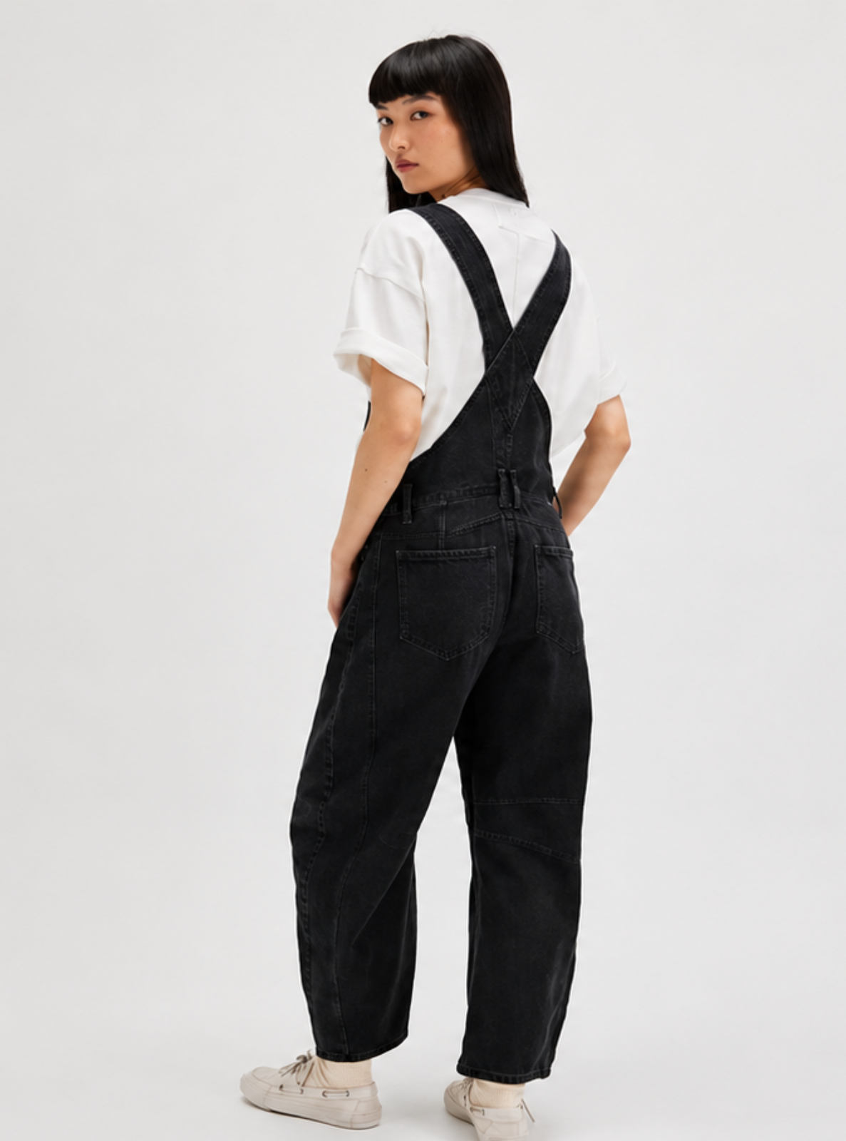 A model wearing black denim overall pants with a white t-shirt, shown from the back against a plain light gray studio background.