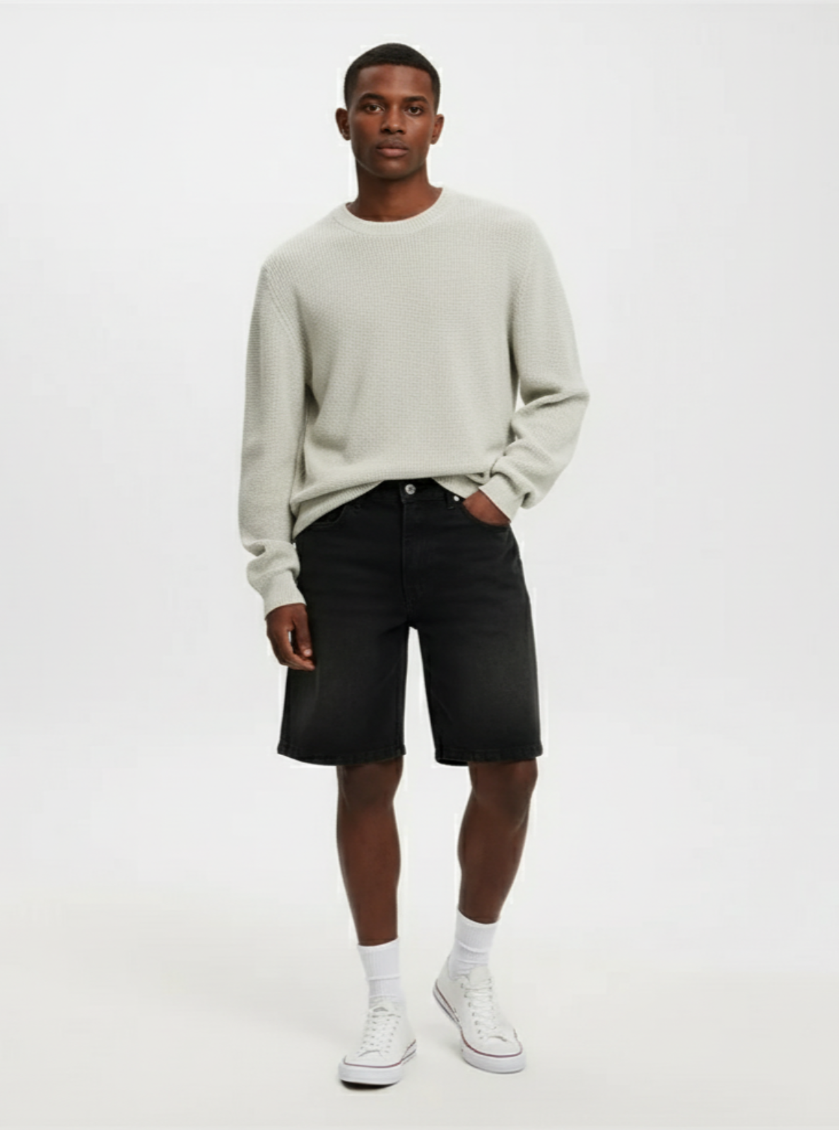 Man wearing a light gray sweater, black shorts, and white sneakers on a white background